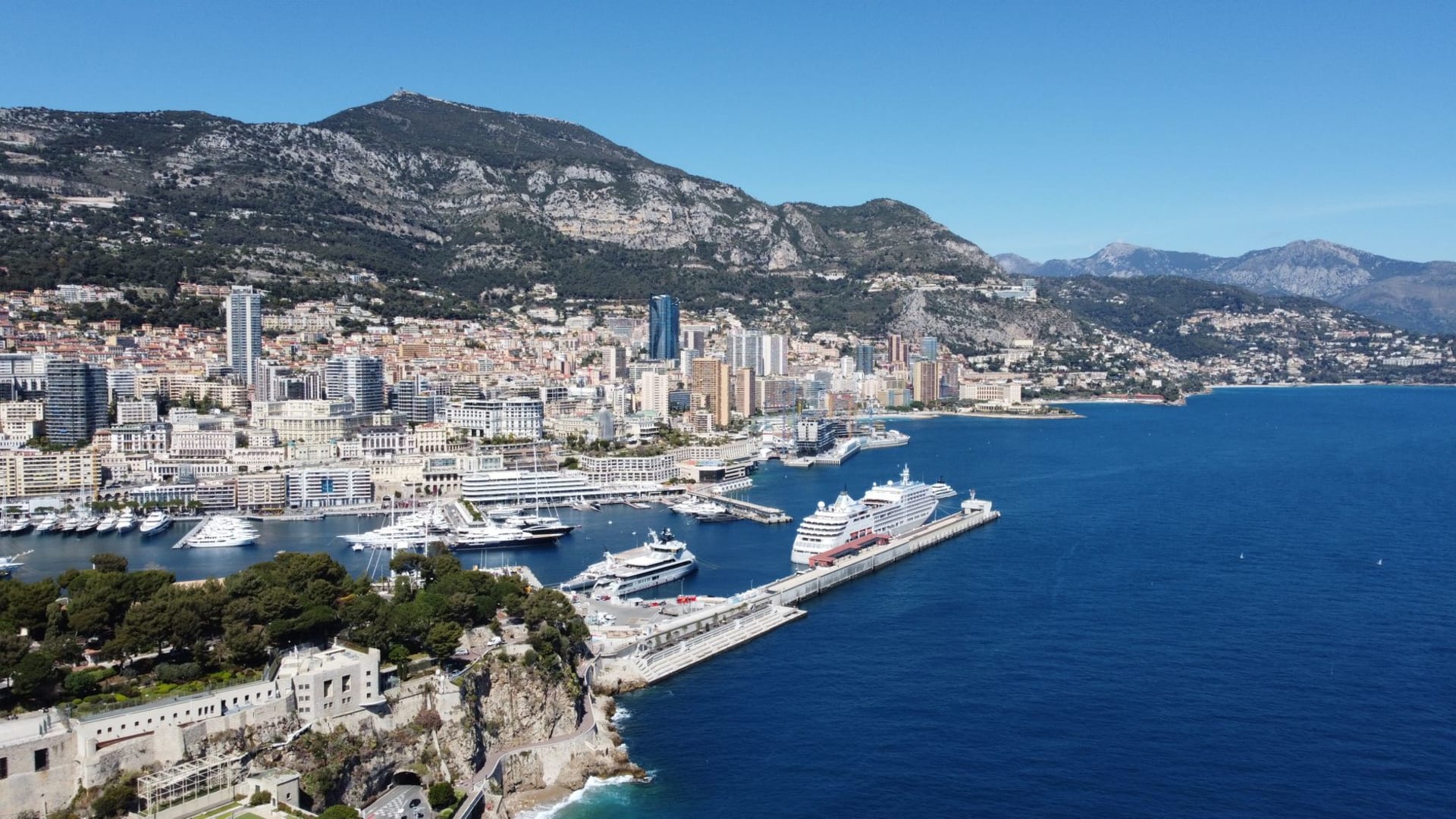 Aerial view of Monaco's harbor packed with superyachts, the dense cityscape climbing the hillside above the blue Mediterranean
