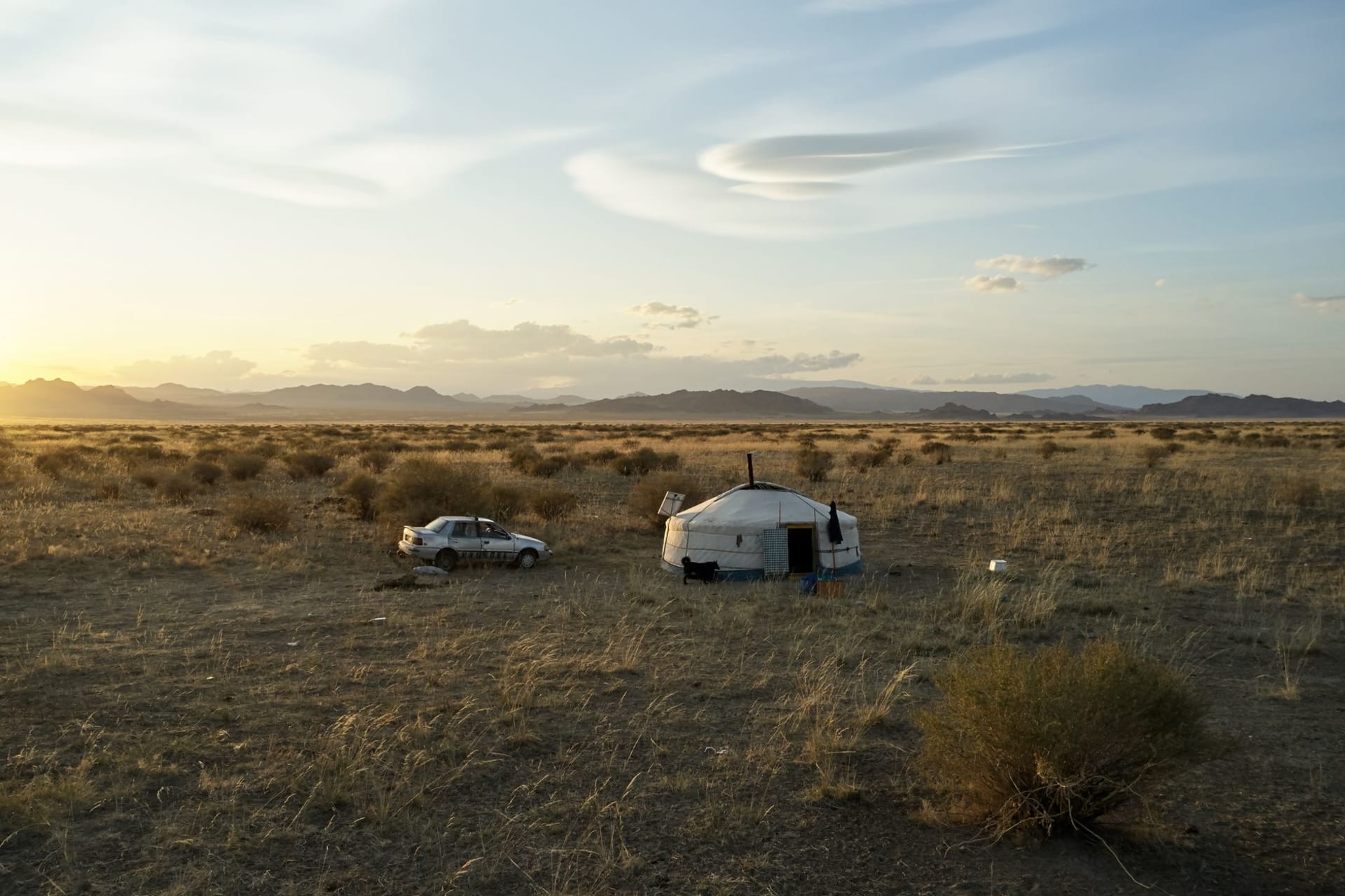 A white traditional ger and a traveler's car parked on open Mongolia steppe terrain at golden sunset
