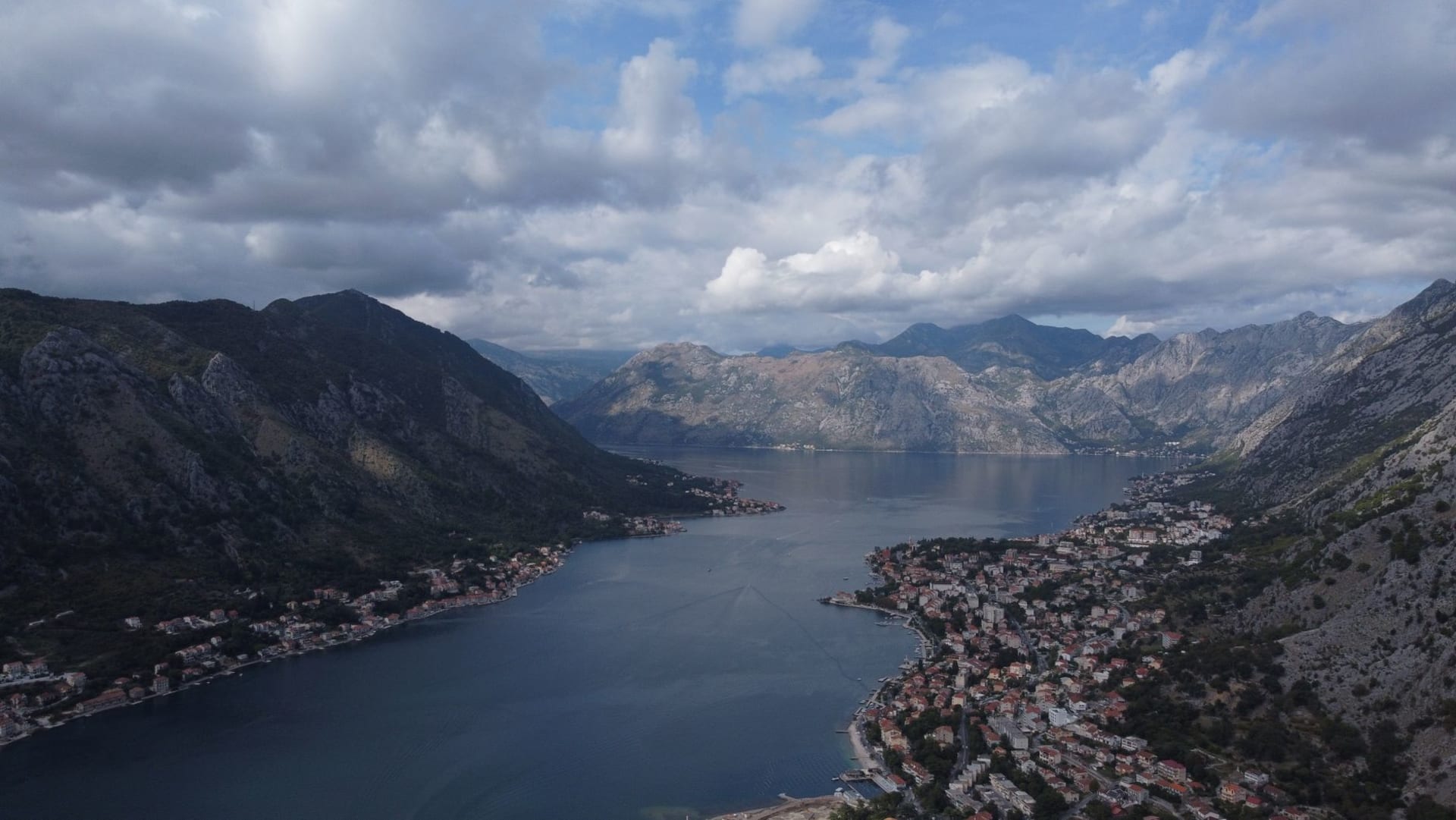 Aerial view of Kotor Bay with its steep mountain walls plunging into dark blue water, a patchwork of terracotta rooftops visible along the shoreline
