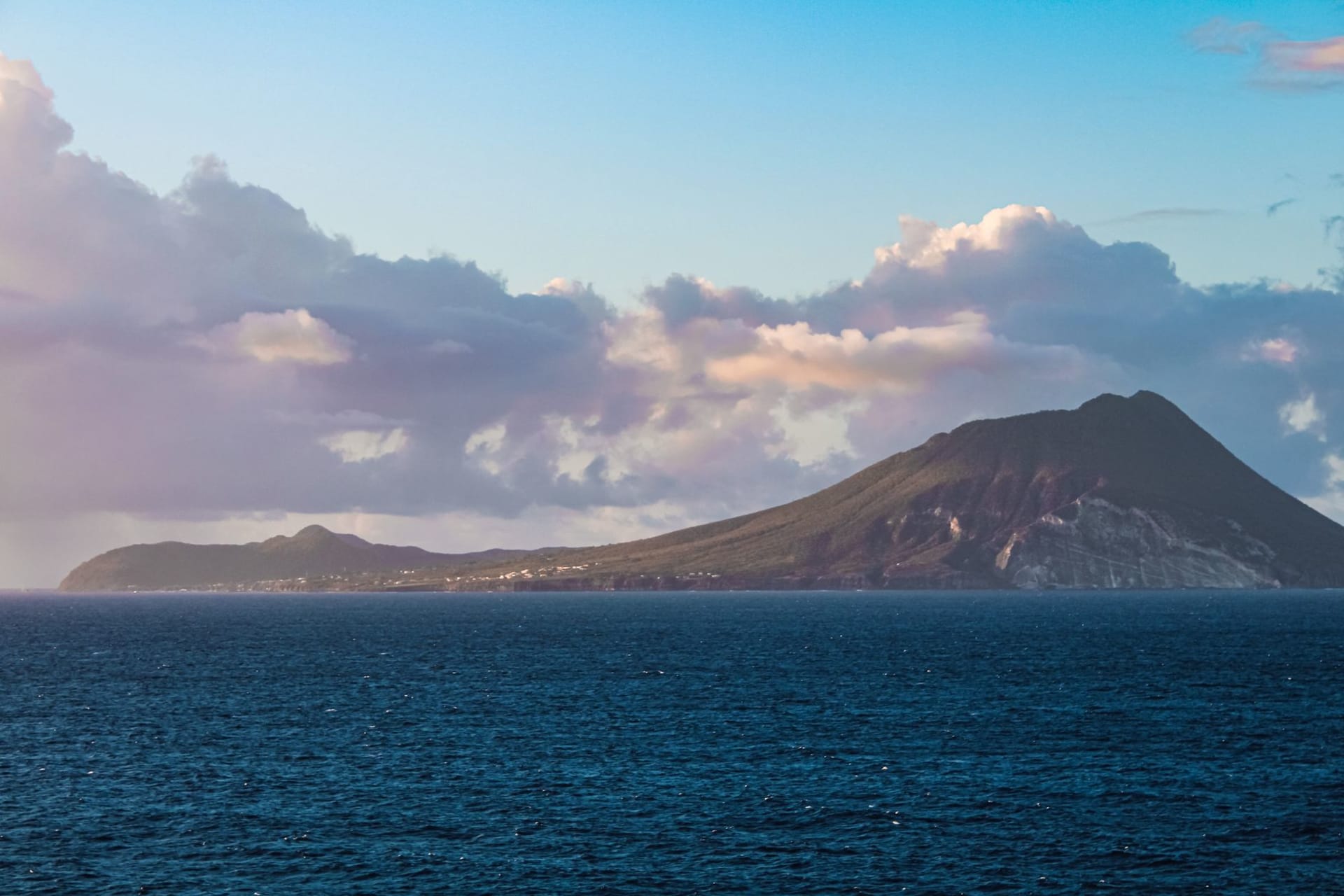Volcanic island rising steeply from a calm turquoise sea, forested slopes meeting the water's edge under a wide sky