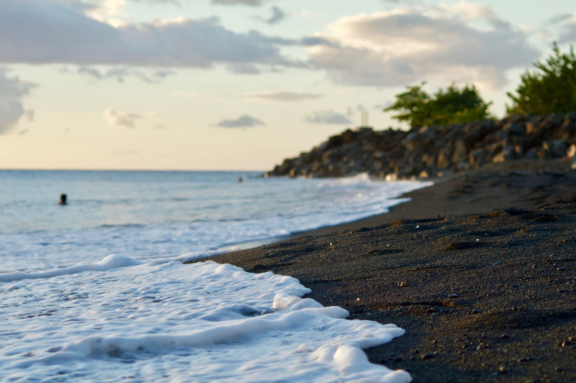 Woodlands Beach Montserrat — dark volcanic sand curving along the shoreline, clear turquoise water, forested hillside behind under an afternoon sky