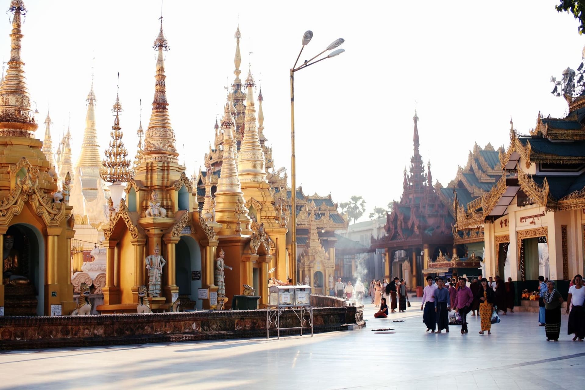 The gilded spire of Shwedagon Pagoda rising above Yangon's crumbling colonial facades at golden hour, the pagoda's surface catching the last light while the street below fills with smoke from tea stall braziers