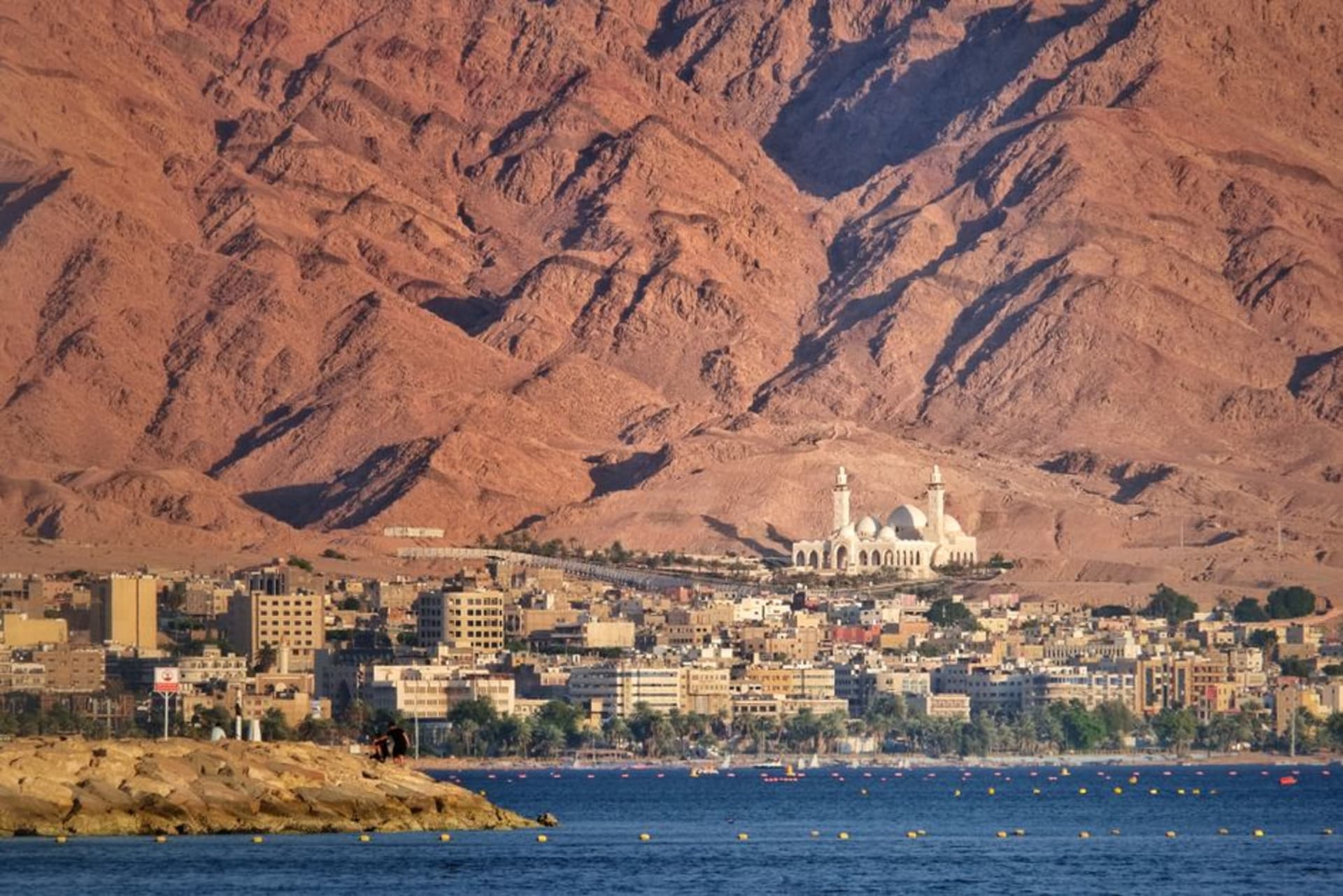 The waterfront of Aqaba at sunset, palm trees along the corniche and the Gulf of Aqaba turning gold, mountains of Sinai visible across the water