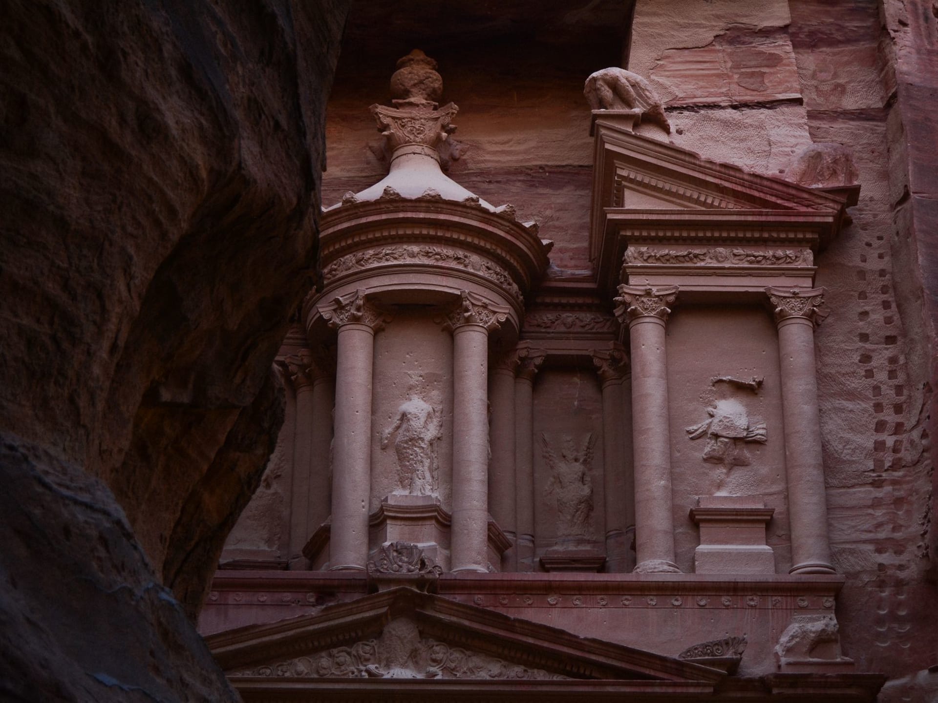 A towering rose-red sandstone facade carved into a Jordanian desert canyon wall, lit by warm afternoon light