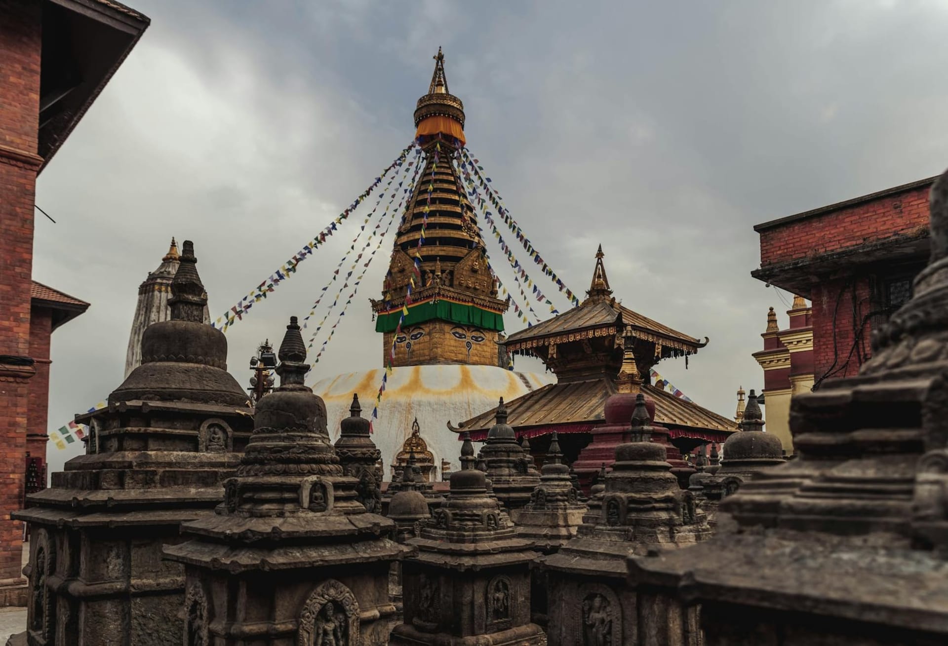 The golden spire of Boudhanath stupa rising above a ring of white-washed walls, butter-yellow prayer flags fanning outward into a hazy Kathmandu sky, with pigeons and monks moving through the courtyard below.