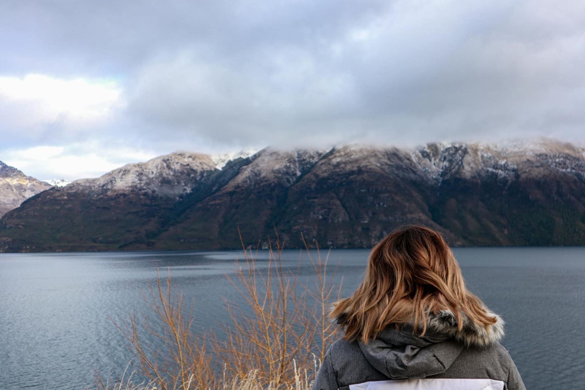 Snow-dusted peaks of the Remarkables reflected in the still blue surface of Lake Wakatipu at dusk, with the lights of Queenstown beginning to glow along the far shore