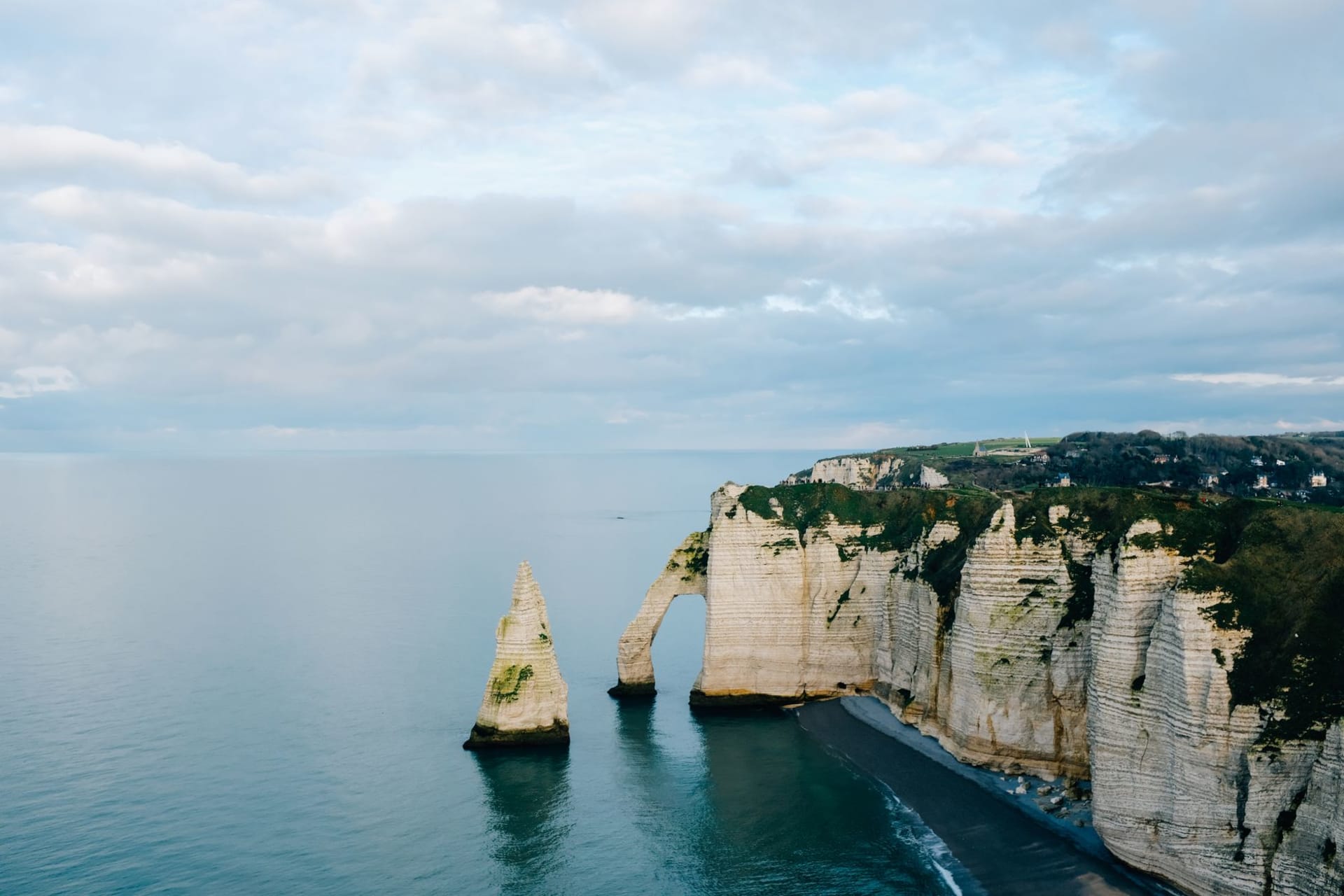 Rocky chalk cliffs covered with green moss rising above the calm grey Atlantic ocean under an overcast sky in Normandy, France