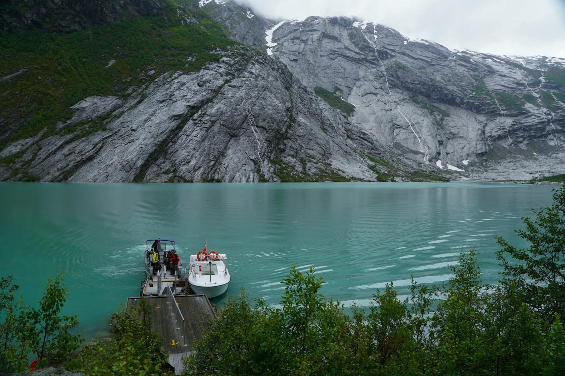 White and pink apple blossoms in full bloom along the banks of Hardangerfjord, with snow-capped mountains rising steeply behind the water and a traditional red farmhouse half-hidden among the trees.