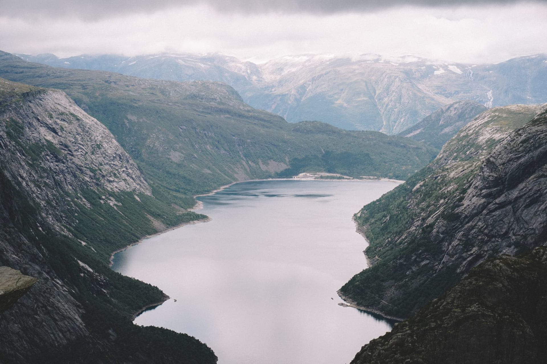 Trolltunga rock formation jutting horizontally over the void, one hiker standing on the tip, Ringedalsvatnet lake far below, mountains all around