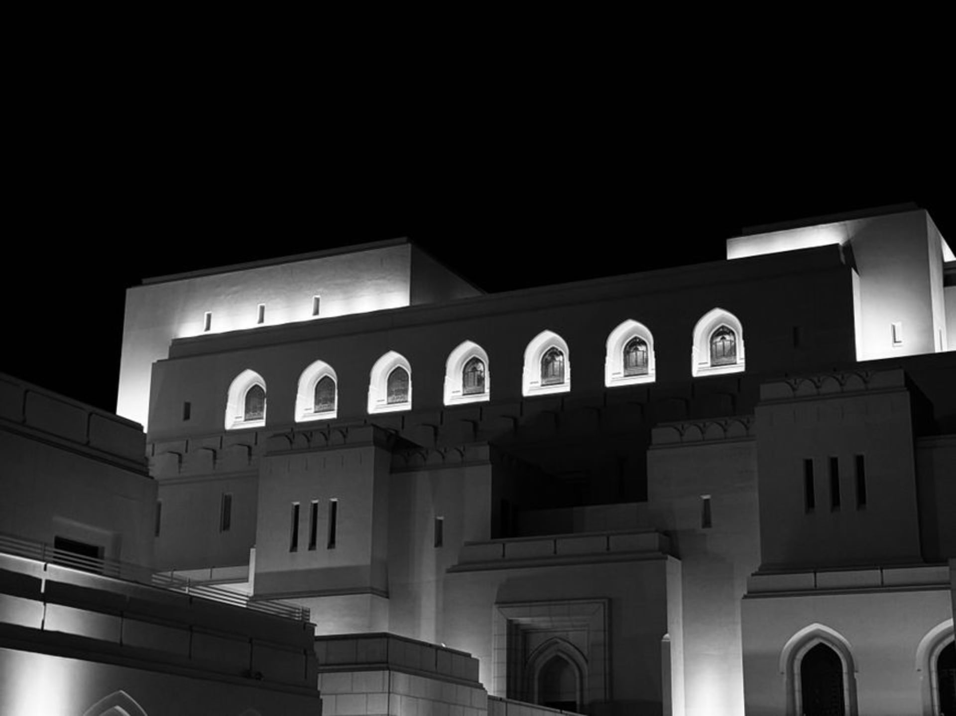 La fachada de arenisca tallada y la columnata arqueada de la Ópera Real de Mascate brillando bajo las luces nocturnas, con jardines cuidados en primer plano.