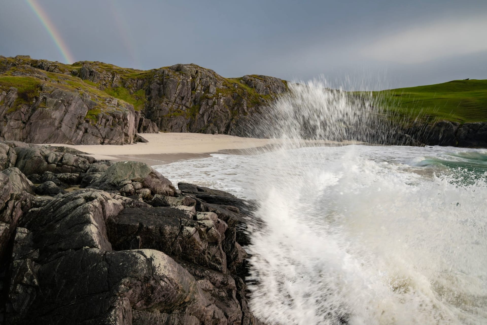 Dramatic waves crashing against the Scottish coastline with a rainbow arcing over the grey Atlantic sky