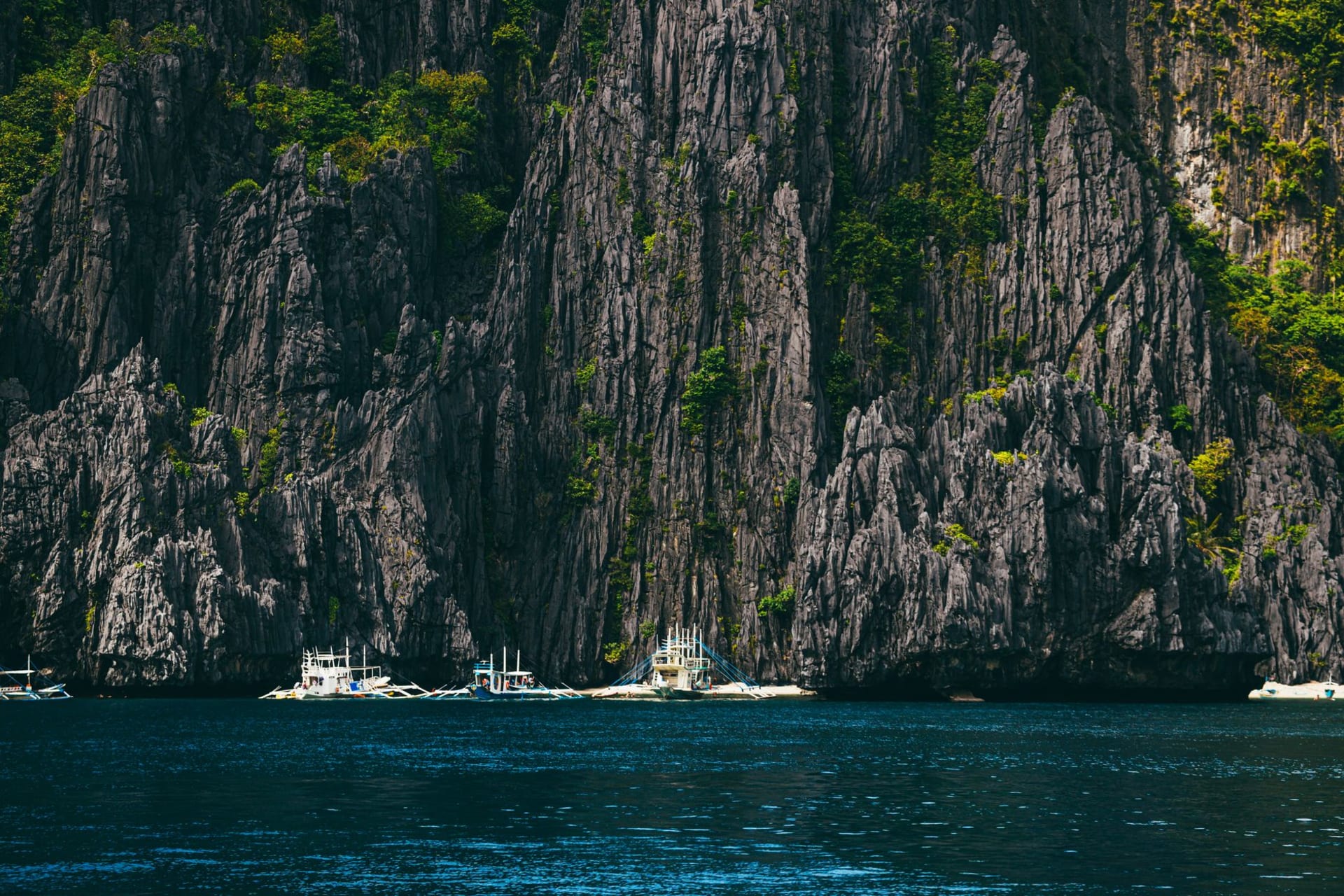 White outrigger bangka boats anchored beneath towering dark limestone karst cliffs in El Nido, Palawan