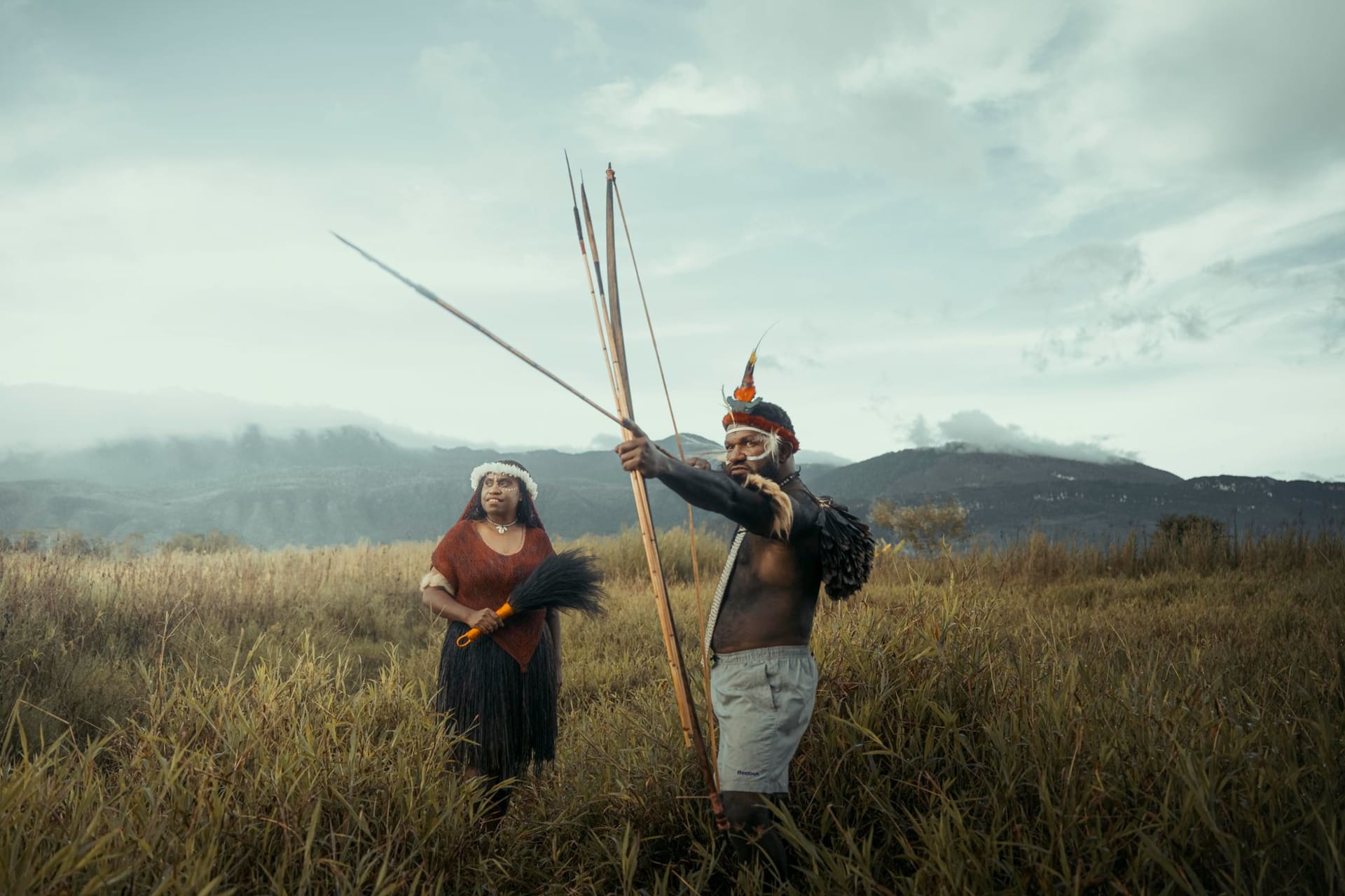 Two indigenous men in traditional dress holding ceremonial spears, standing in a vast green highland landscape with mountains behind them