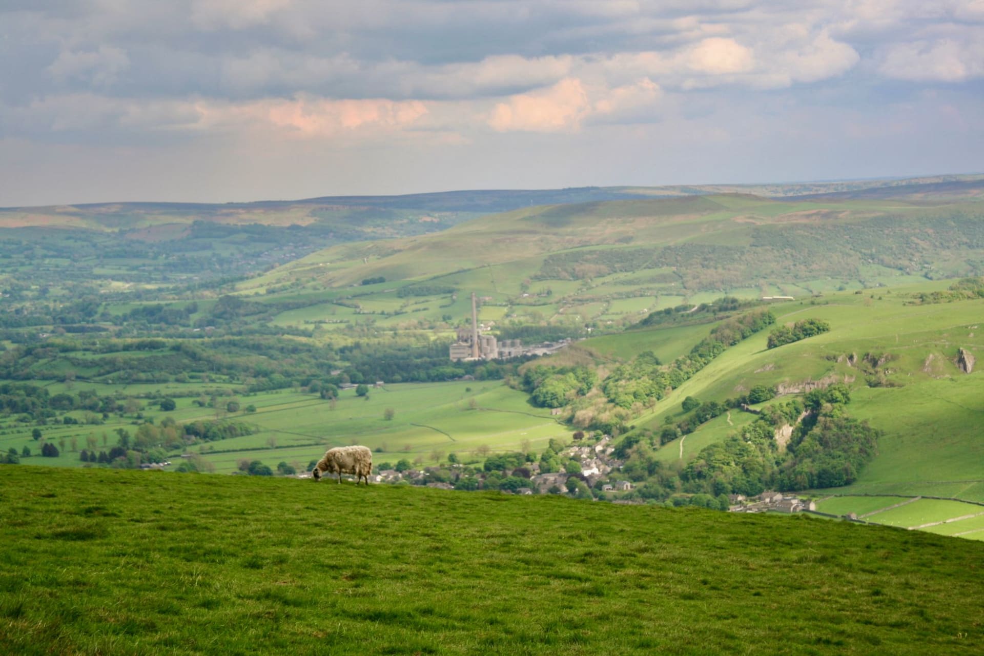 Hathersage village from the hillside above, the church tower visible among trees with Stanage Edge rising on the moor behind in afternoon light