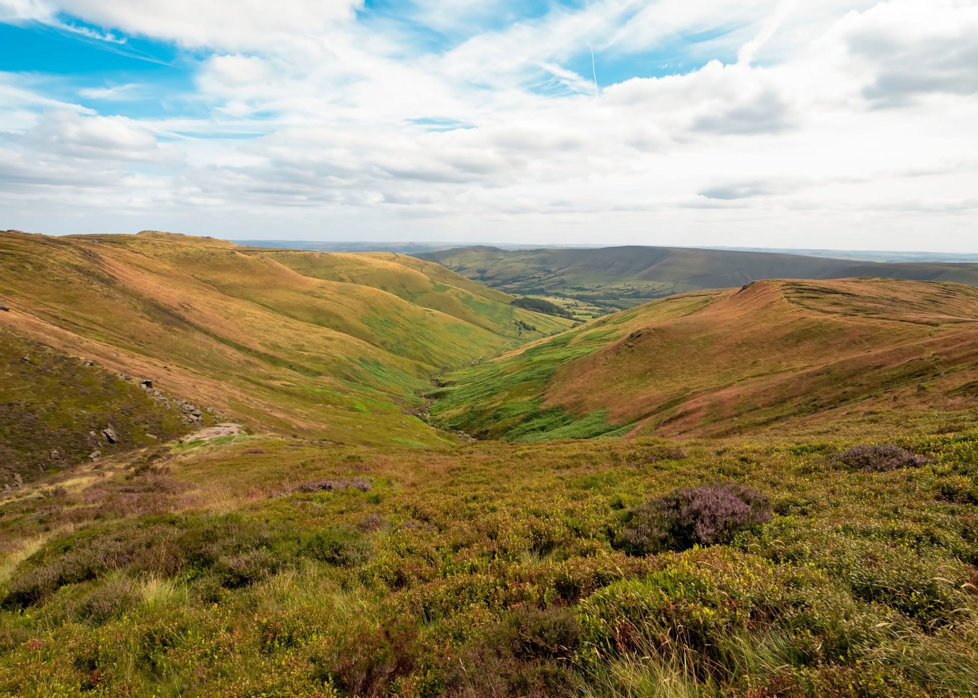 A sweeping moorland valley with heather-covered slopes rolling toward a green dale under a cloud-streaked sky in the Peak District