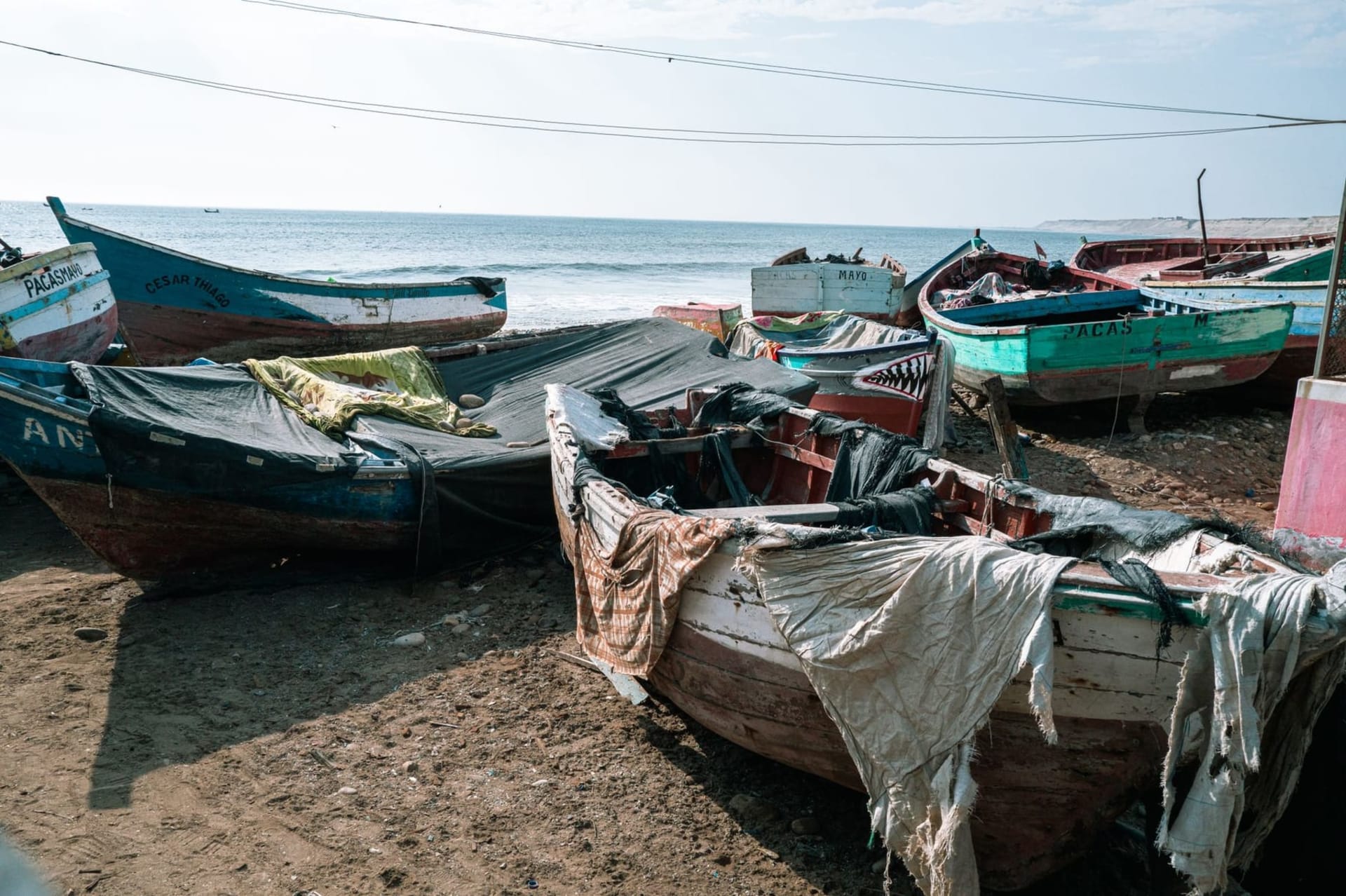 Golden late-afternoon light over Mancora's main beach, where wooden fishing boats rest at the waterline and surfers carry boards across pale sand toward small breaking waves.