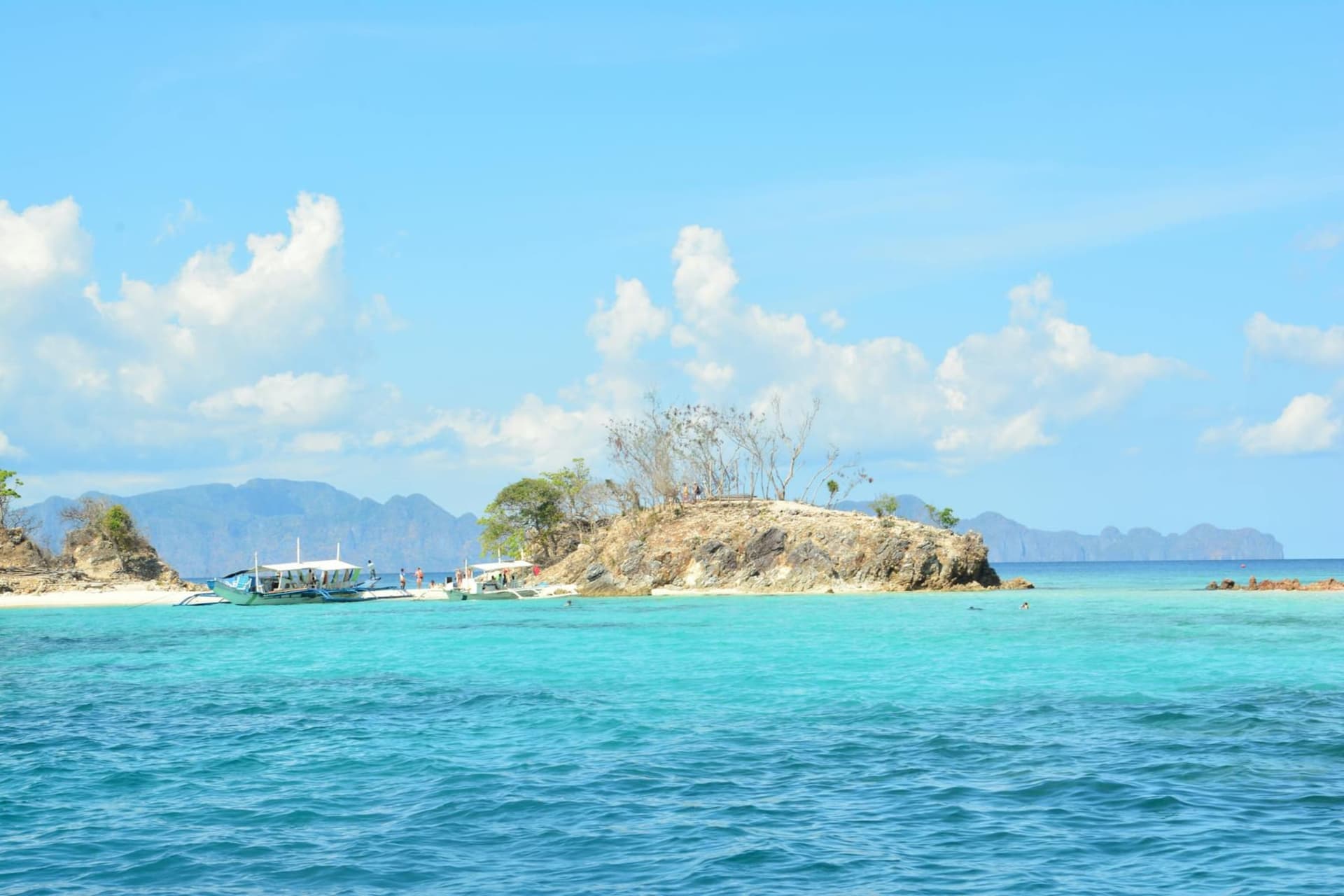 A diver silhouetted above the barnacled hull of a Japanese WWII shipwreck in the turquoise shallows off Sangat Island, limestone karst cliffs rising from the sea in the background.