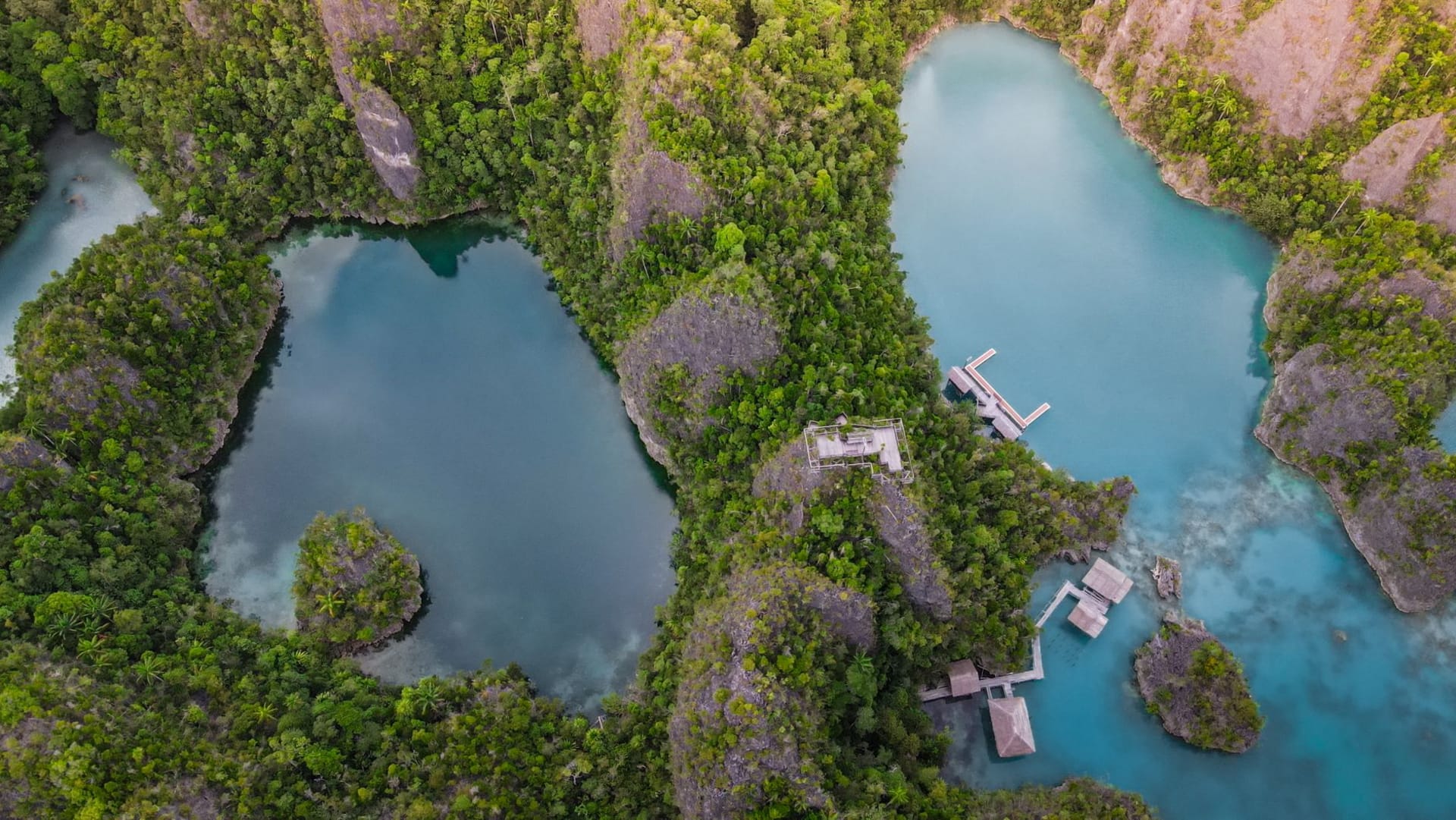 Gam Island's forested ridge rising steeply above a calm turquoise bay, simple wooden homestay bungalows on stilts along the shore and a longboat moored in the shallows