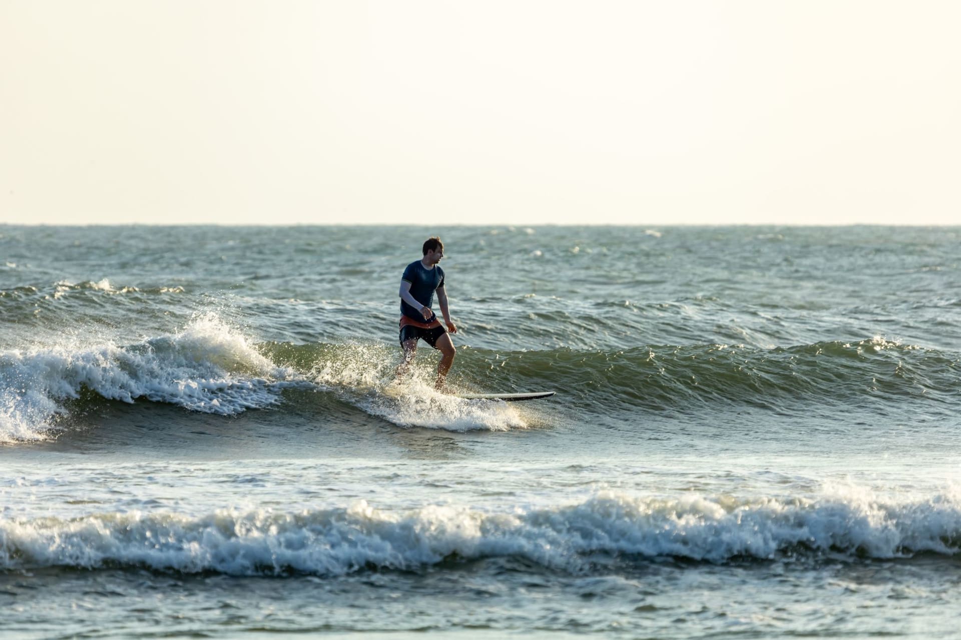 A surfer on a long right-hand wave at Saint-Leu point break, the volcanic coastline and green hills visible behind, the Indian Ocean glittering in late afternoon sun