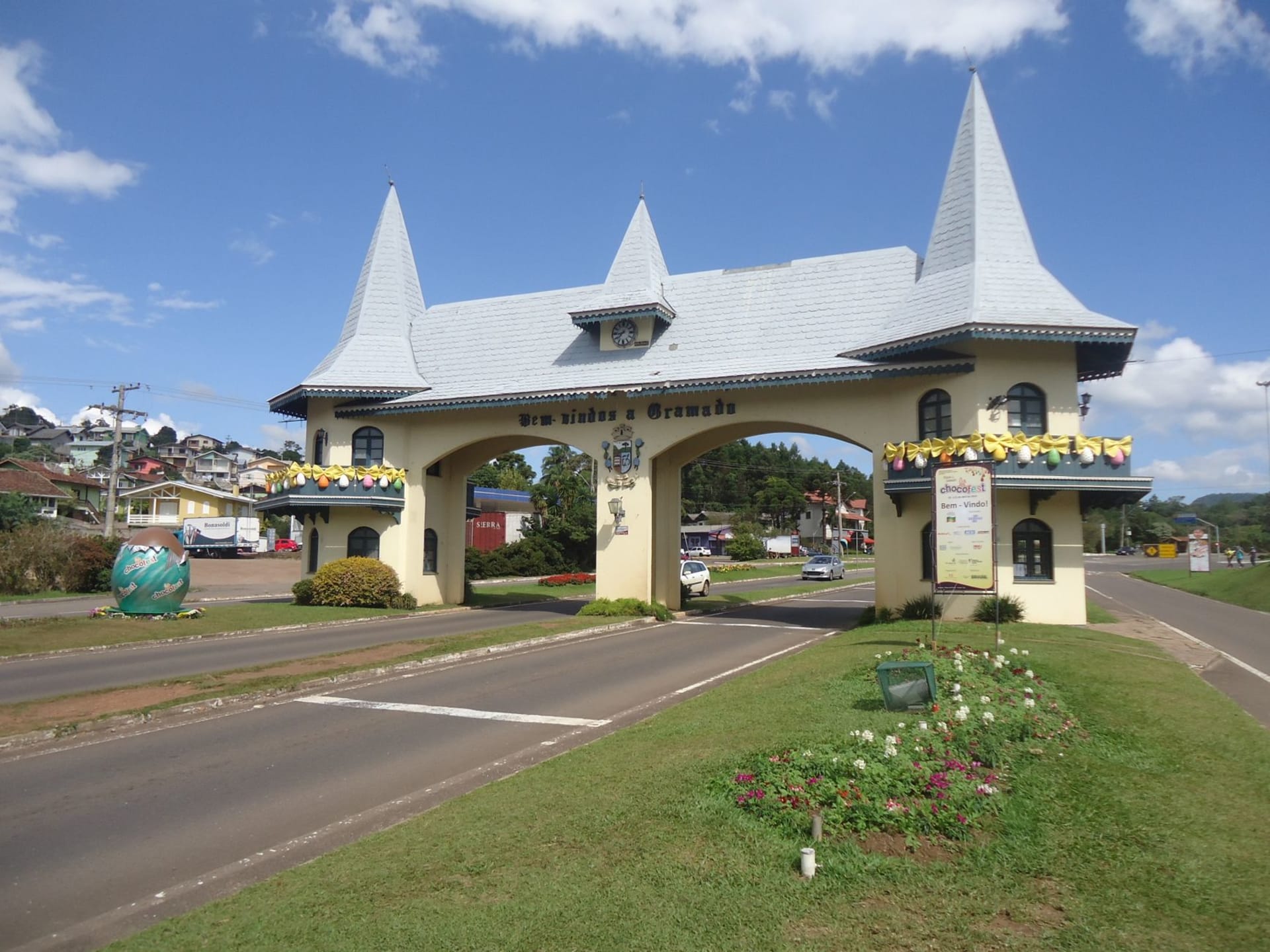 Ornate entrance arch of Gramado lit by warm afternoon light, surrounded by alpine-style architecture and lush green hills