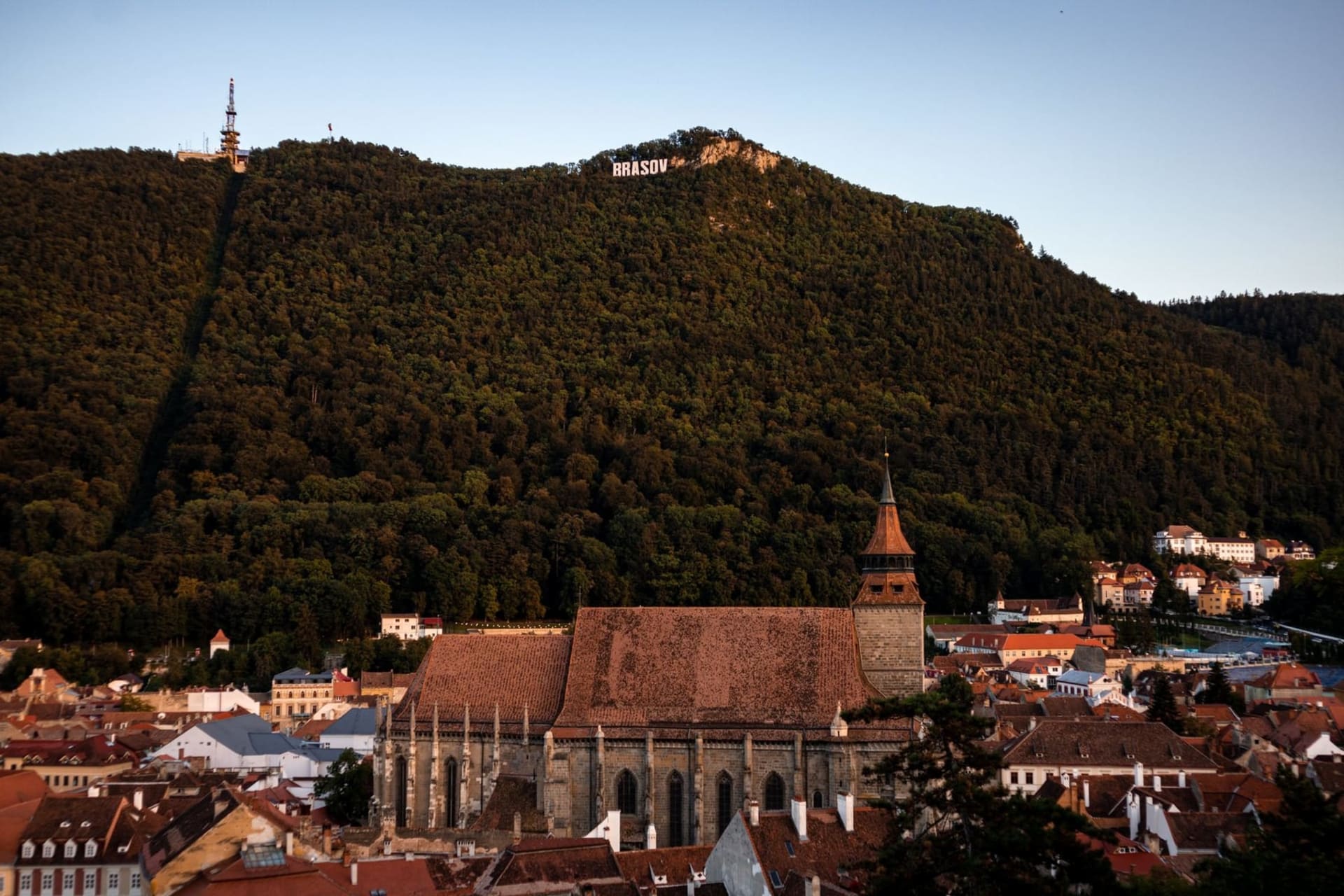 Panoramic view of Brasov's red-roofed old town from Tampa Mountain, with the Black Church's dark Gothic spire rising above the medieval square and forested Carpathian peaks filling the horizon