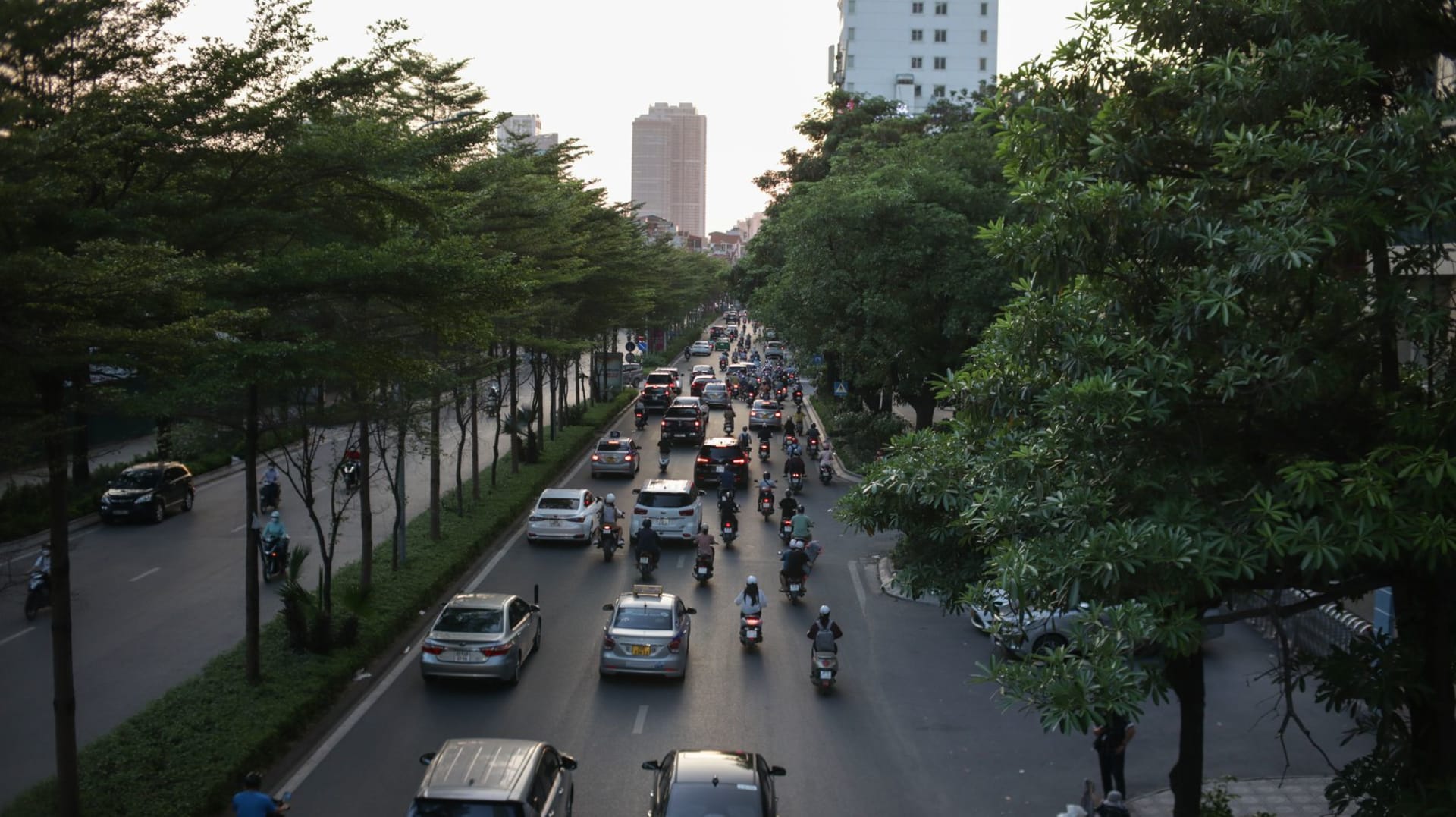 Ouagadougou's central avenue at dusk, motorbike traffic flowing in both directions under orange streetlights, food vendors set up along the road
