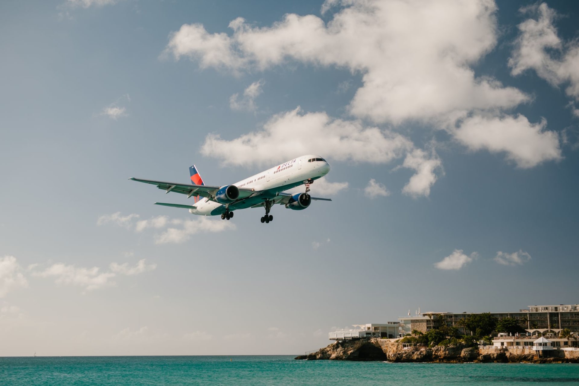 A commercial aircraft descending low over a turquoise Caribbean bay, just above the beach at Maho, with clear blue skies and a resort in the background