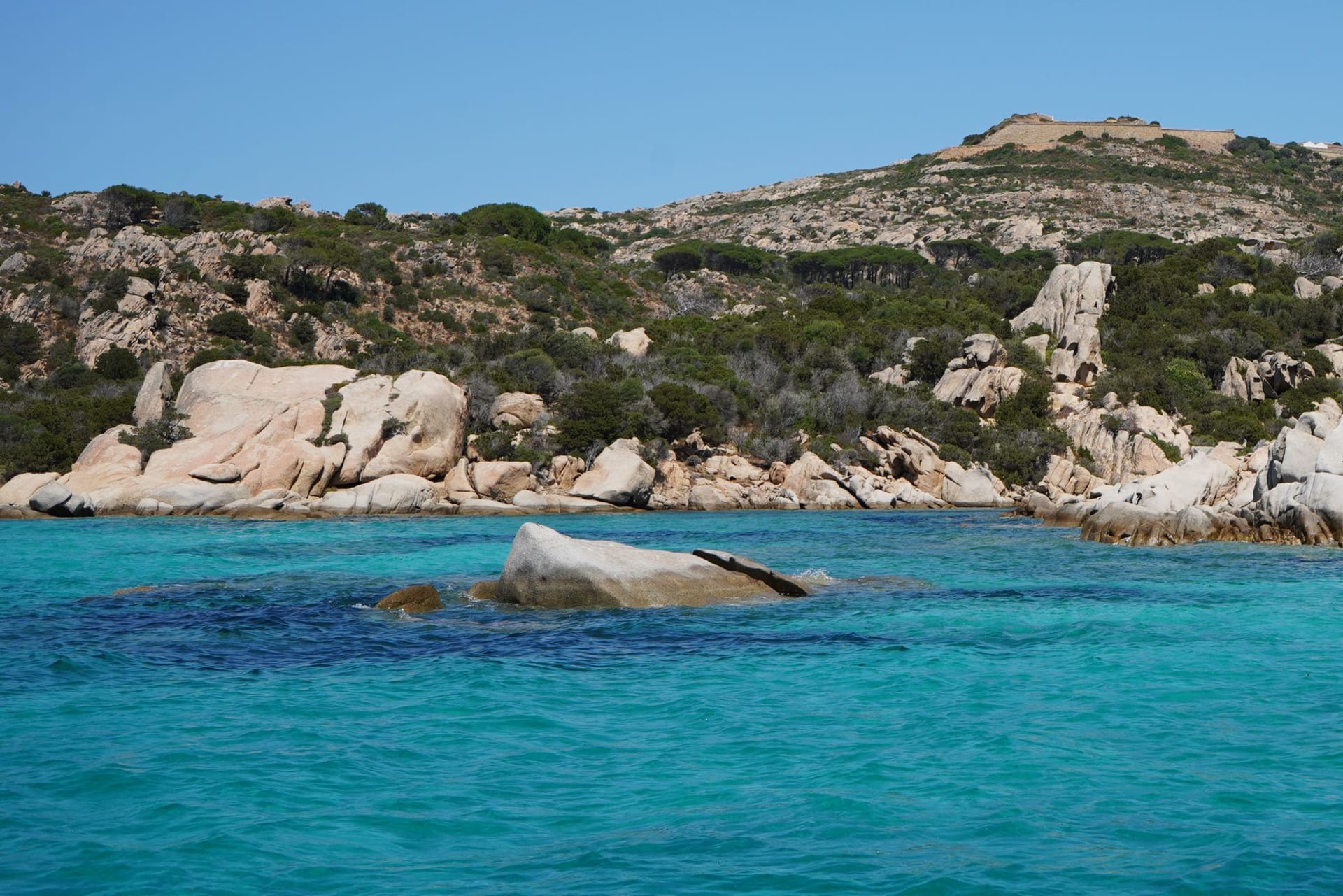 Agua turquesa cristalina sobre arena blanca de granito en Spiaggia del Principe con grandes rocas de granito rosado en primer plano