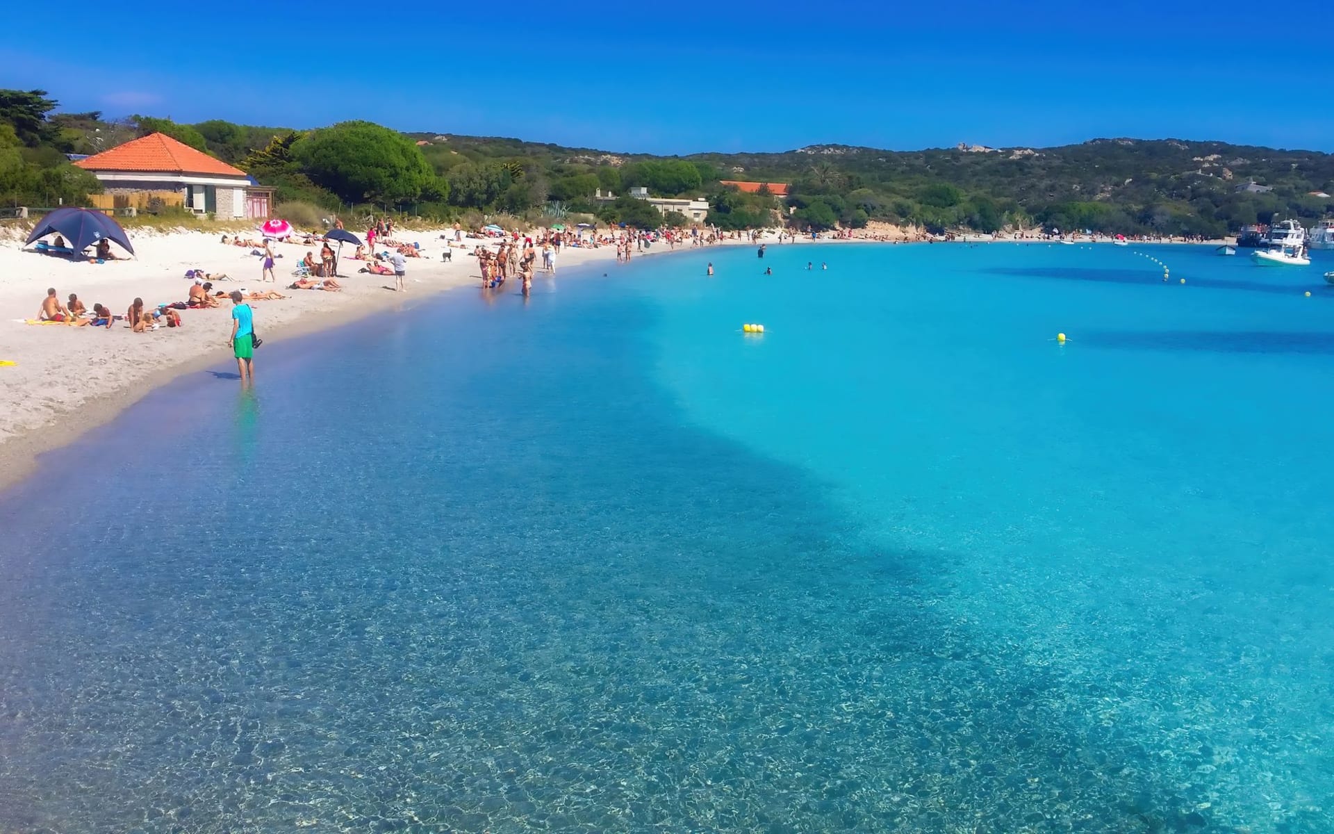 Suaves rocas de granito rosado del archipiélago de La Maddalena emergiendo de un agua de claridad turquesa imposible, con una segunda isla visible en el horizonte bajo un cielo azul