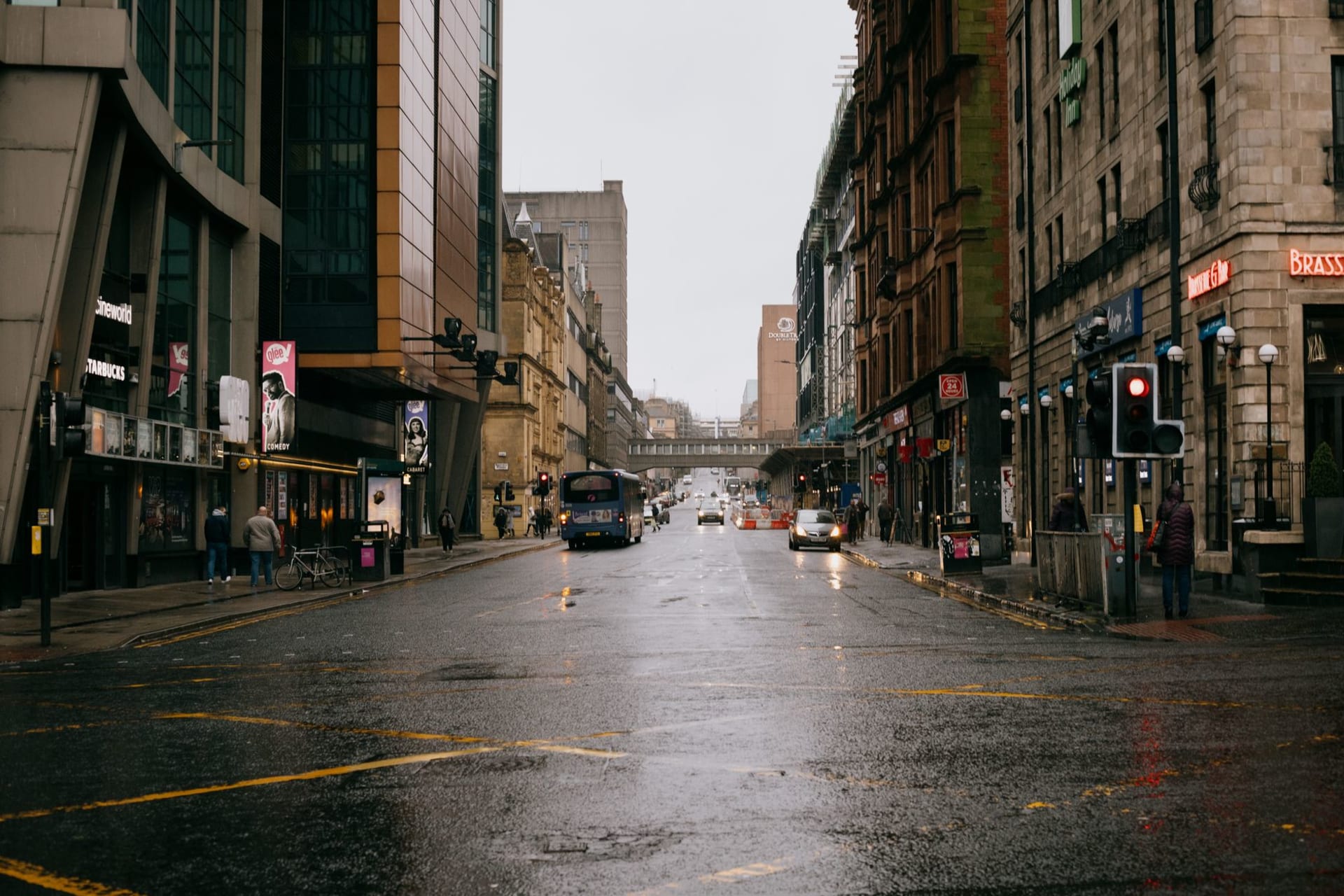 Glasgow's George Square at dusk under heavy grey cloud, blackened Victorian sandstone facades and the floodlit City Chambers reflecting in wet paving stones