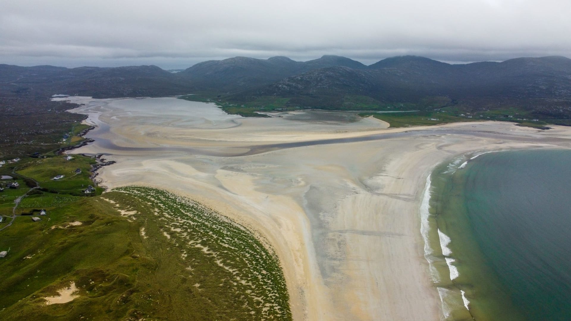 Luskentyre beach with shallow turquoise water, white shell-sand and dark gneiss mountains looming behind under a cloud-streaked Atlantic sky