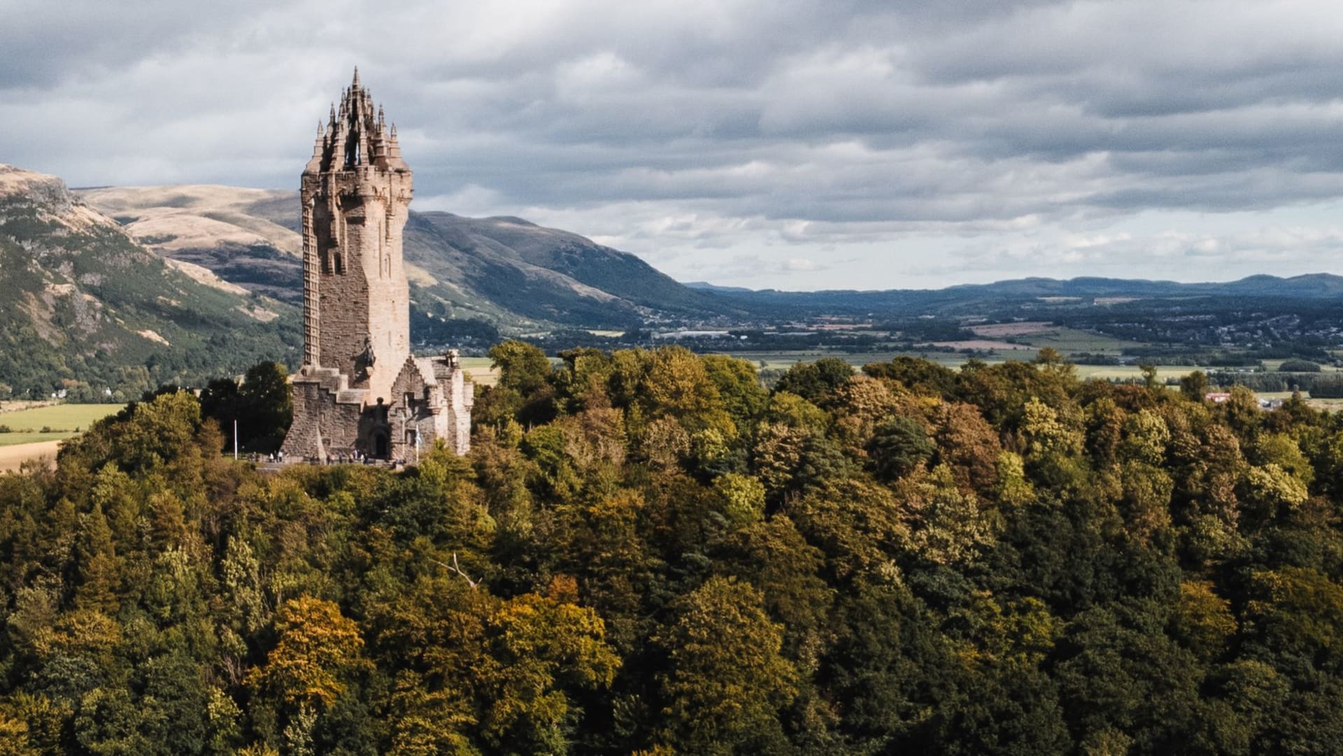 El castillo de Stirling encaramado en su roca volcánica bajo un cielo tormentoso, con la torre del Monumento a Wallace visible sobre su colina arbolada en la distancia media