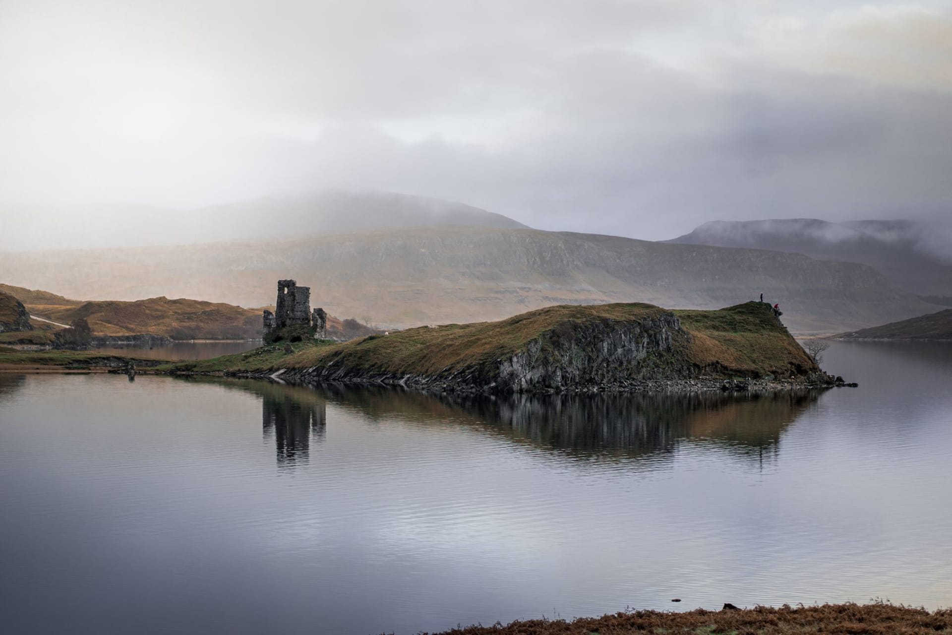 Ardvreck Castle ruins reflected on the still surface of Loch Assynt, wrapped in low mist with mountains dissolving into grey sky