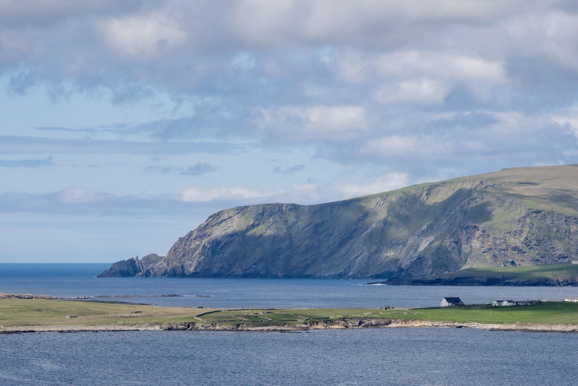 Fair Isle from the air showing the green crofting land, dramatic cliffs on the south end, and the tiny harbour, surrounded on all sides by dark Atlantic water