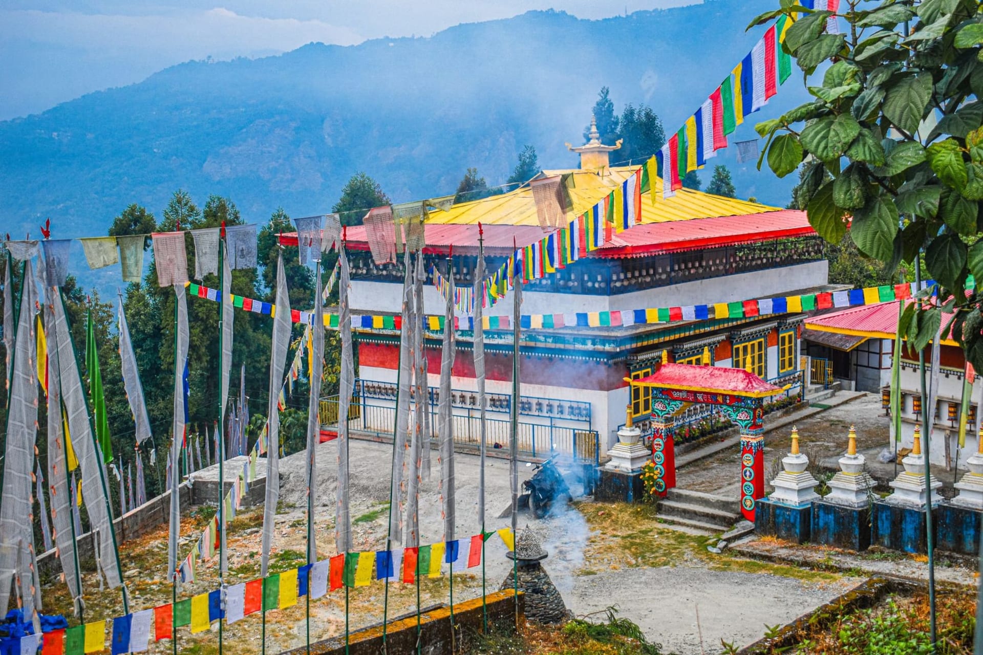 A Buddhist monastery with white walls and red-trimmed windows perched on a green ridge above Pelling, with Kangchenjunga's snow-capped triple summit rising behind it in crisp morning light.