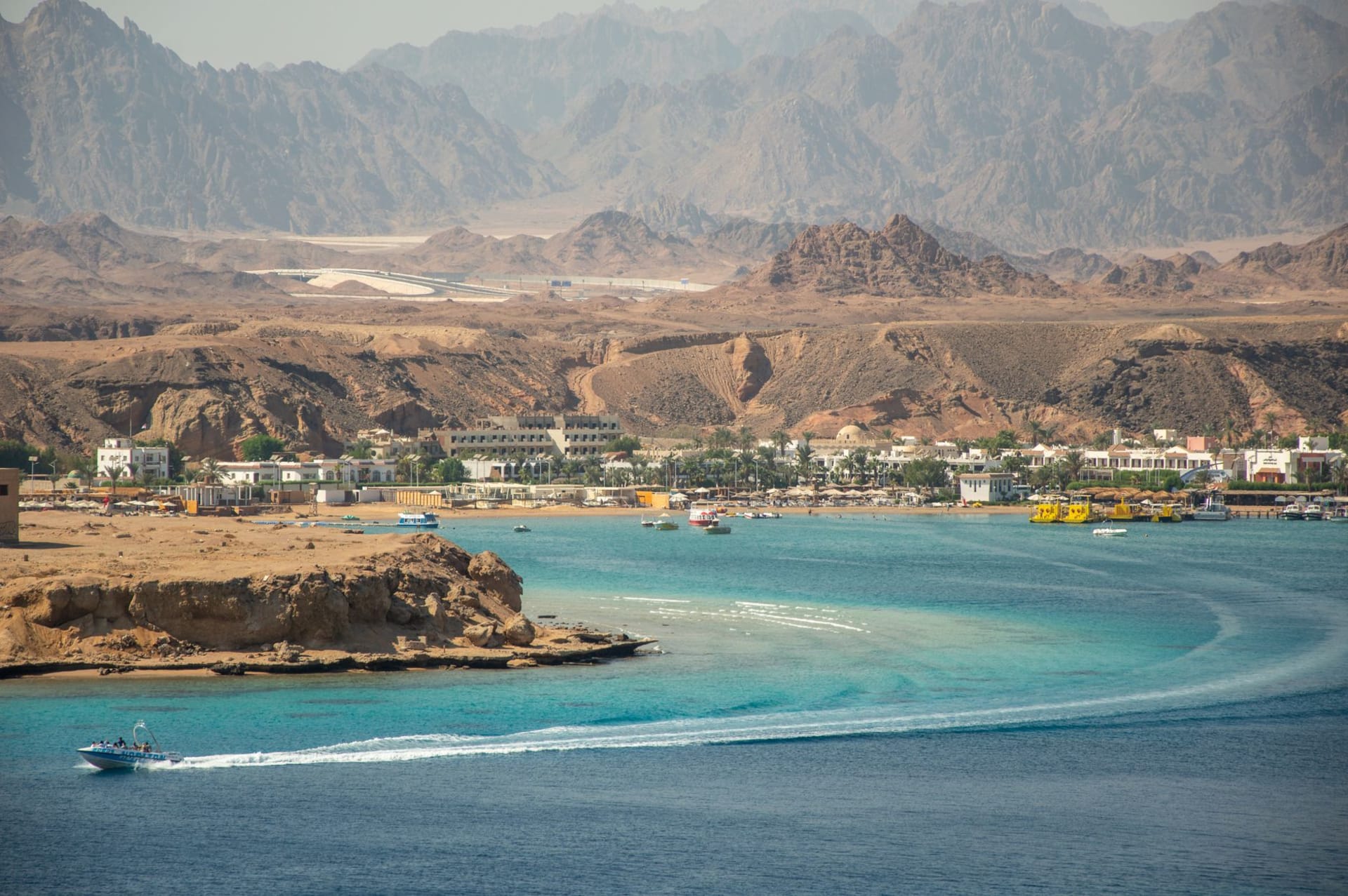 An aerial view of the Blue Hole at Dahab, the circular dark indigo sinkhole set against turquoise shallows with a sandy coral reef fringe, desert cliffs rising behind