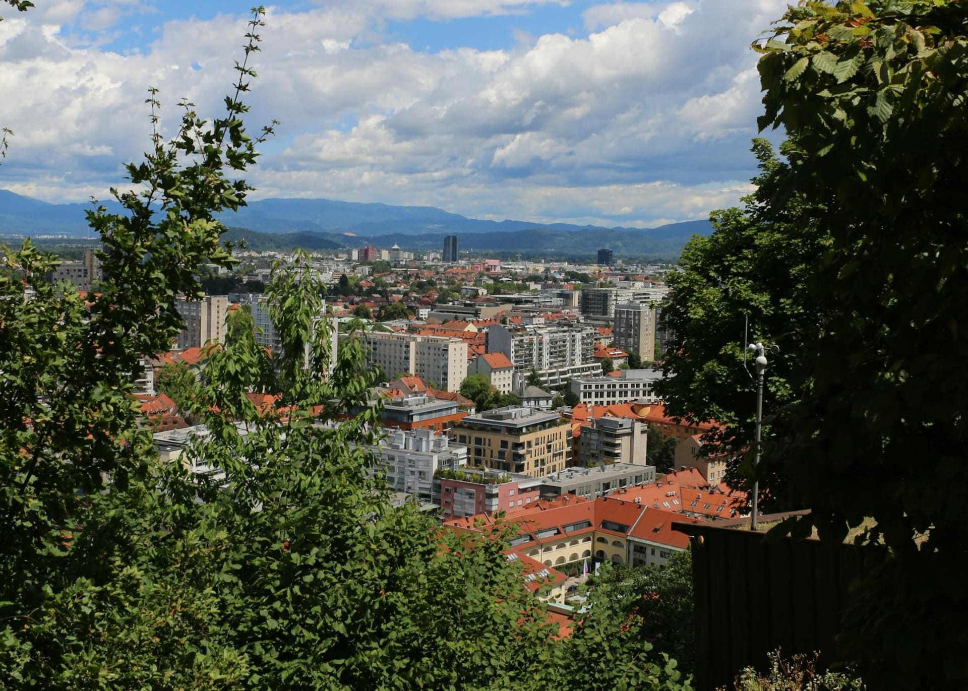 The Ljubljanica river winding beneath Ljubljana Castle on its hilltop, with orange-roofed buildings lining the water and the Tromostovje triple bridge in the foreground on a clear afternoon.