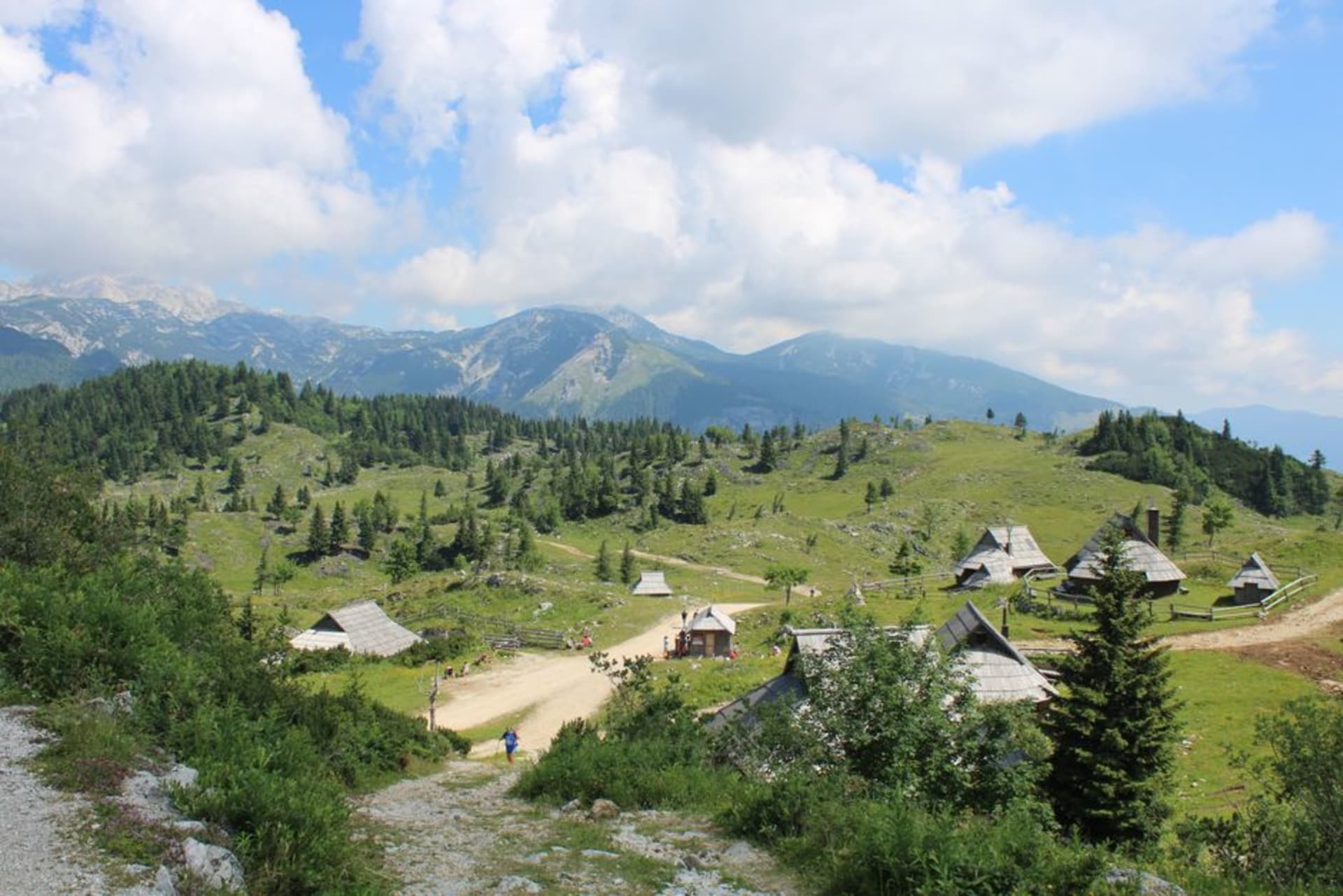 Praderas alpinas onduladas y verdes de Menina Planina bajo un cielo abierto, con cabañas de pastores de madera dispersas y vistas lejanas hacia los Alpes de Kamnik-Savinja