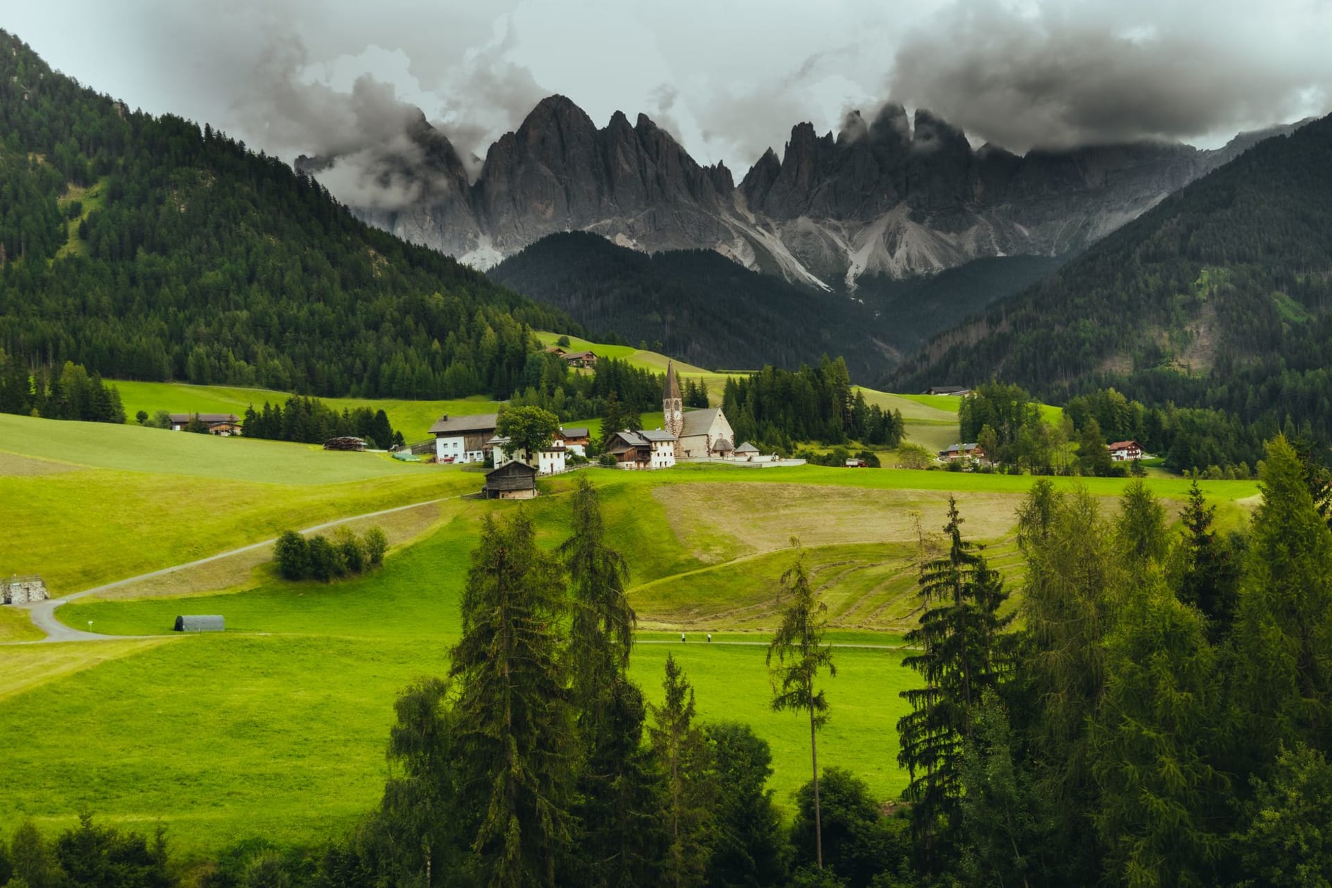 An alpine village of pale stone houses clustered beneath dramatic storm-lit Dolomite peaks and a sweep of deep green meadows