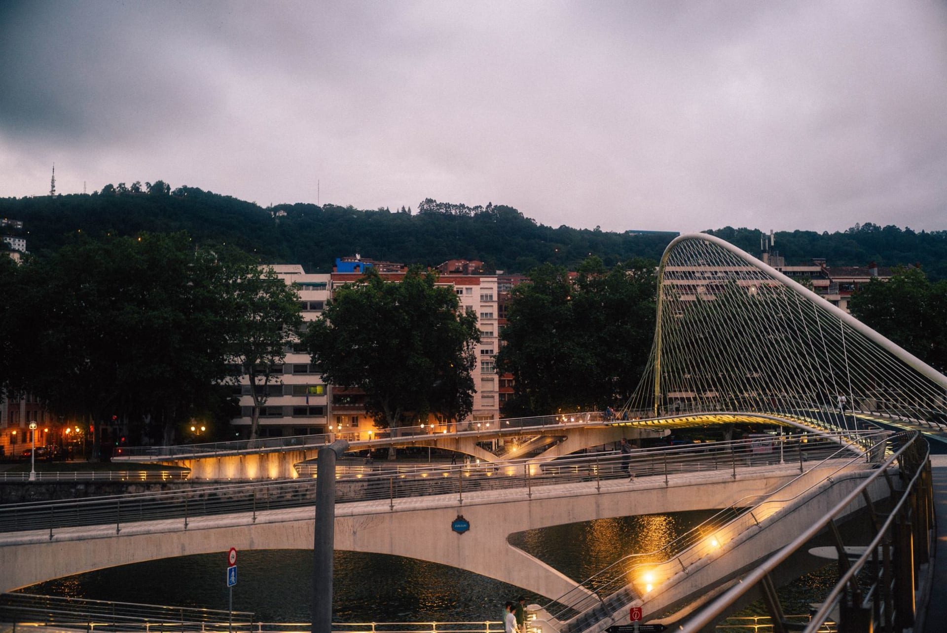 The titanium curves of the Guggenheim Bilbao catching afternoon light along the Nervión river, with the Puente de la Salve bridge arching red in the background