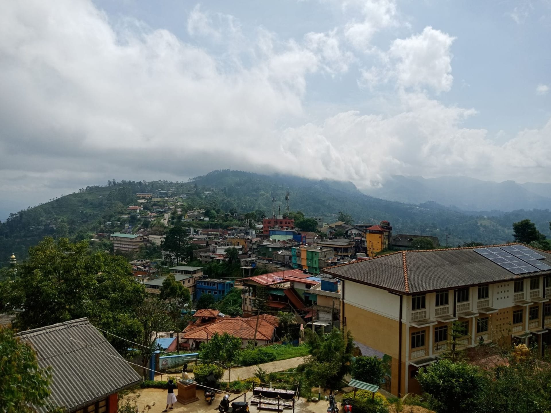 Terraced tea plantations cascading down a steep ridge in Haputale, with low cloud drifting through the valley below and a Tamil tea plucker in a bright sari working the rows in the foreground.