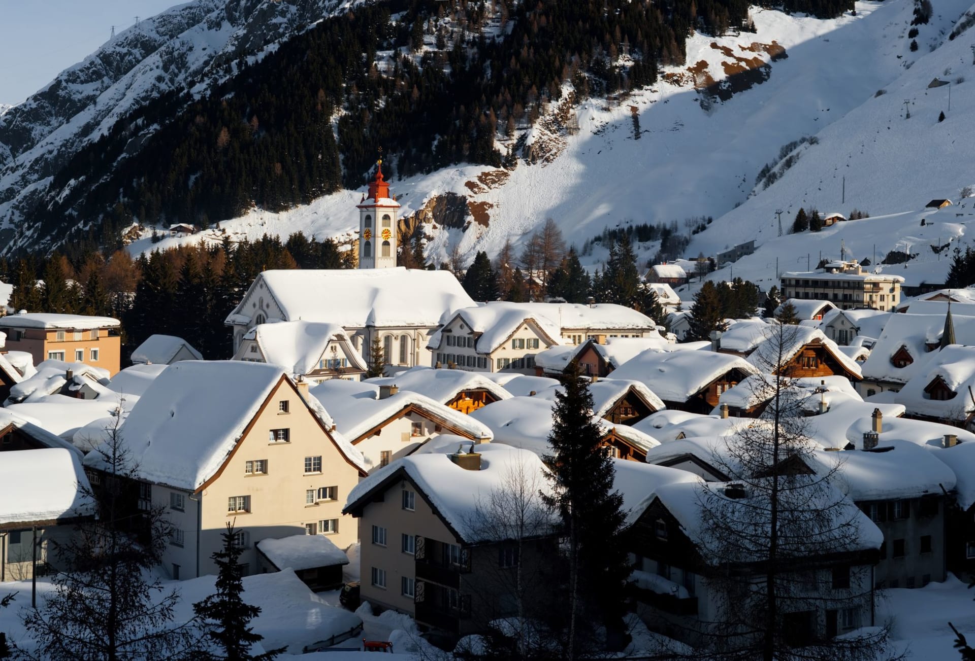 Le village d'Andermatt avec son église en pierre grise et ses toits serrés au cœur de la large vallée d'Urseren, le Gemsstock s'élevant abruptement au-dessus dans la neige hivernale et la dure lumière alpine