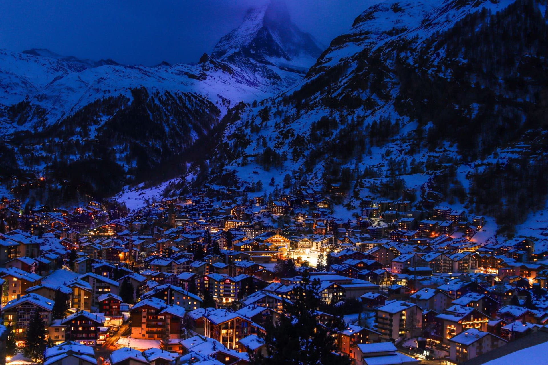 Nighttime view of Zermatt village with illuminated chalet houses and snow-covered alpine peaks in the background