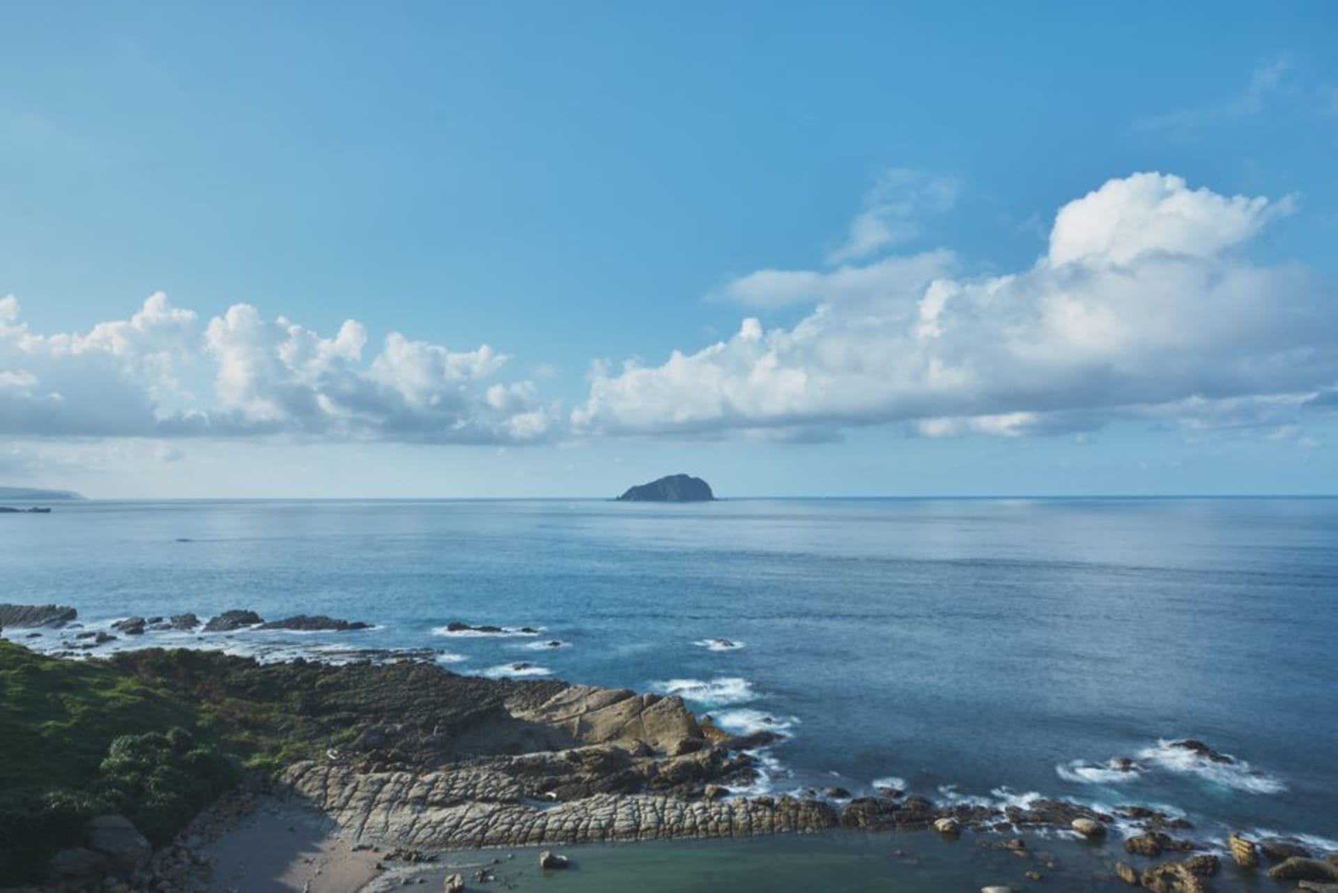 A white lighthouse on a green hill overlooking the narrow sandbar of Cijin Island with the ocean and Kaohsiung's harbor entrance beyond.