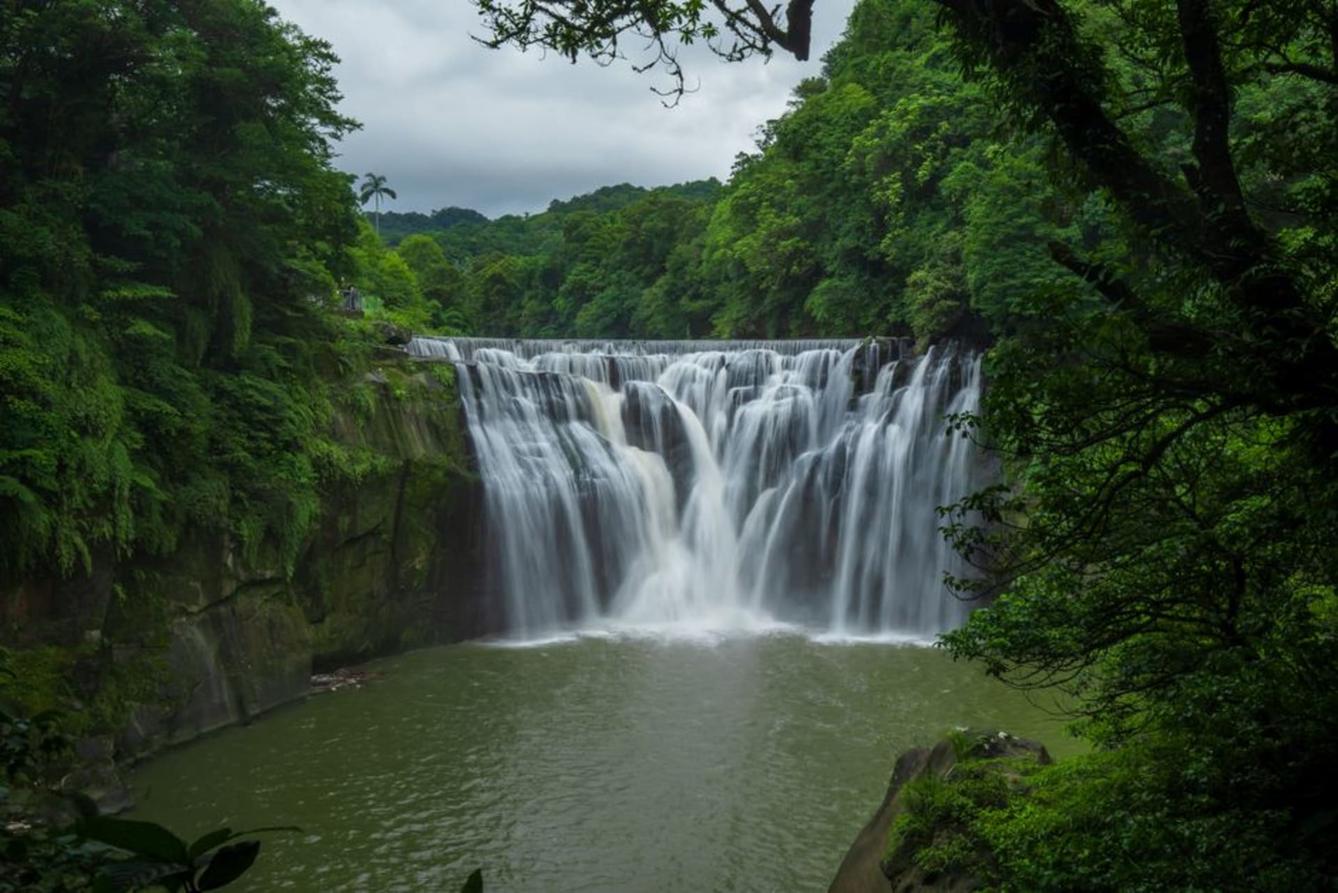 A wide, curtain-like waterfall at Shifen crashing into a turquoise pool surrounded by tropical forest