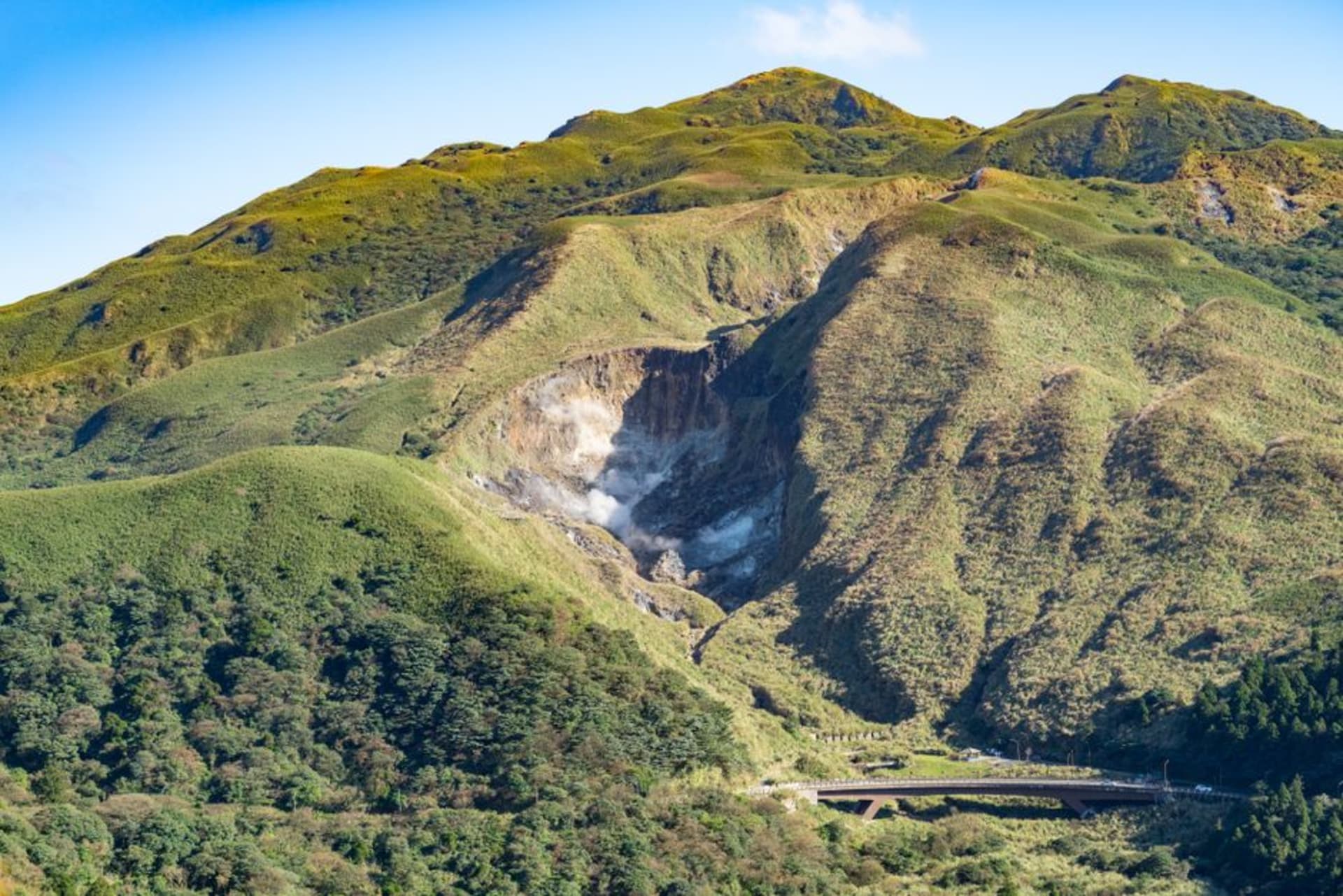 Fumarolas de azufre liberando vapor sobre un paisaje volcánico árido en Xiaoyoukeng, en Yangmingshan, con cordilleras verdes al fondo