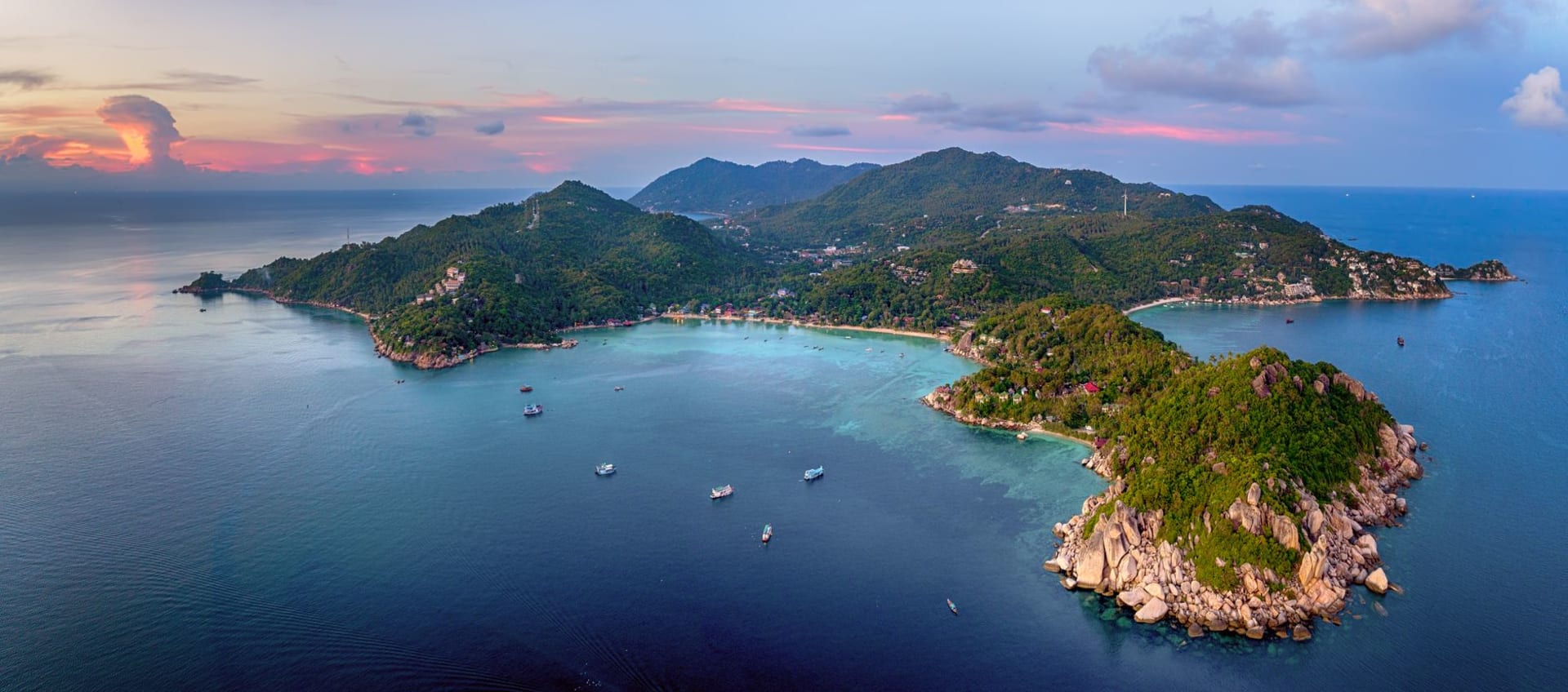 Turquoise shallow water over a coral reef off Koh Tao, Thailand, with a dive boat anchored in the distance and a limestone-forested hillside rising behind the bay.