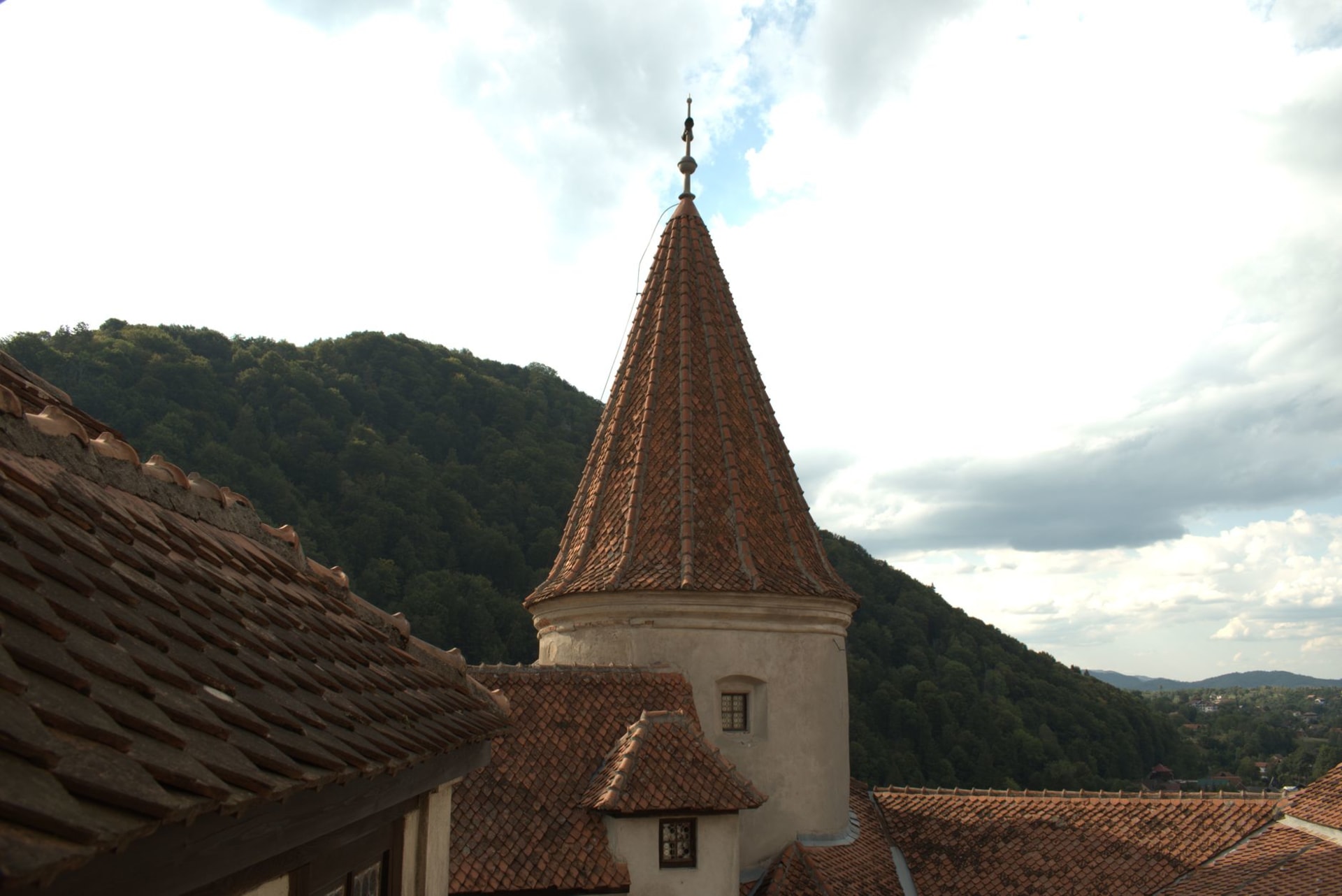 Bran Castle rising from its rocky outcrop against a backdrop of forested Carpathian hills, white towers and terracotta roofs under a dramatic overcast sky