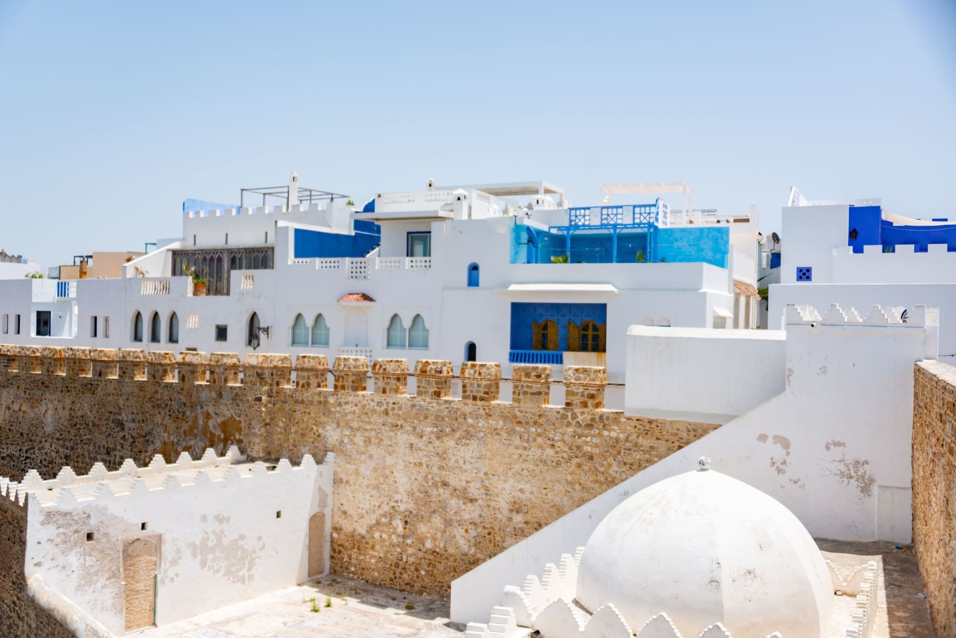 The thick crenellated walls of the Sousse ribat tower rising against a cloudless morning sky, a tiled rooftop and the blue glimmer of the Mediterranean visible in the background