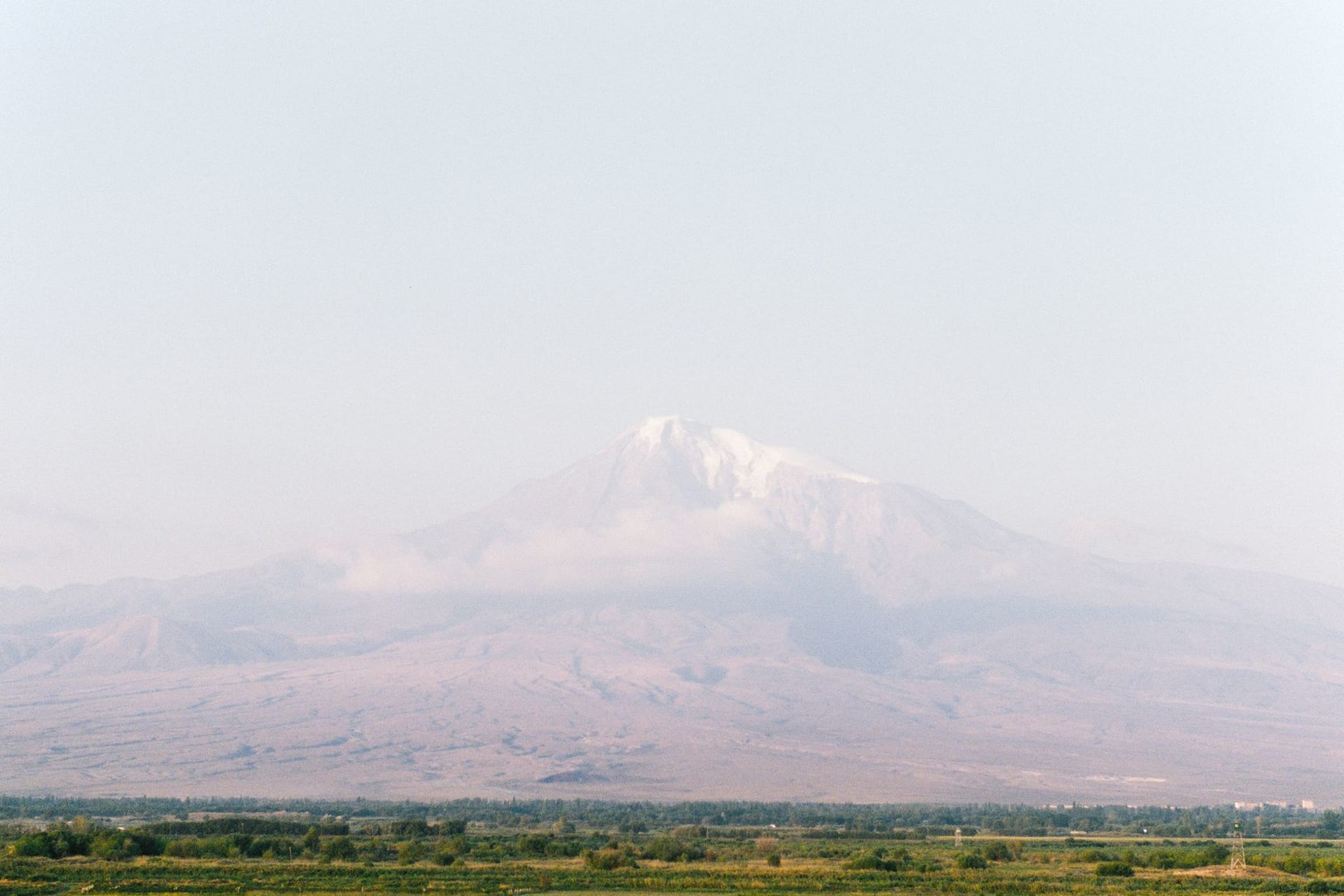 The snow-capped twin-peaked cone of Mount Ararat rising alone above golden plains and a small village in far eastern Turkey under a clear blue sky.